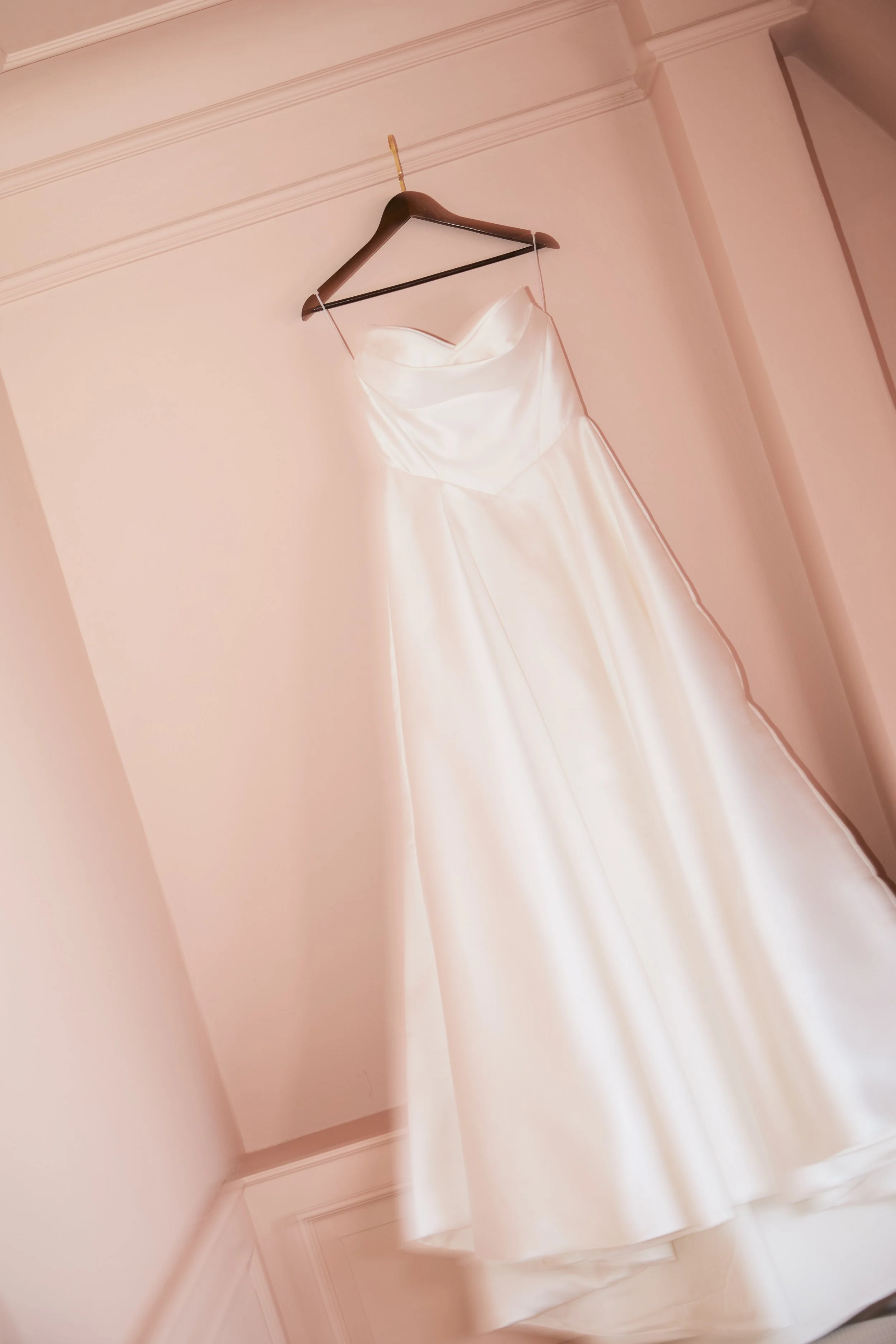 Ein weißes Brautkleid hängt an einem Holzkleiderbügel an einer Wand in einem Raum mit hellrosa Wänden.