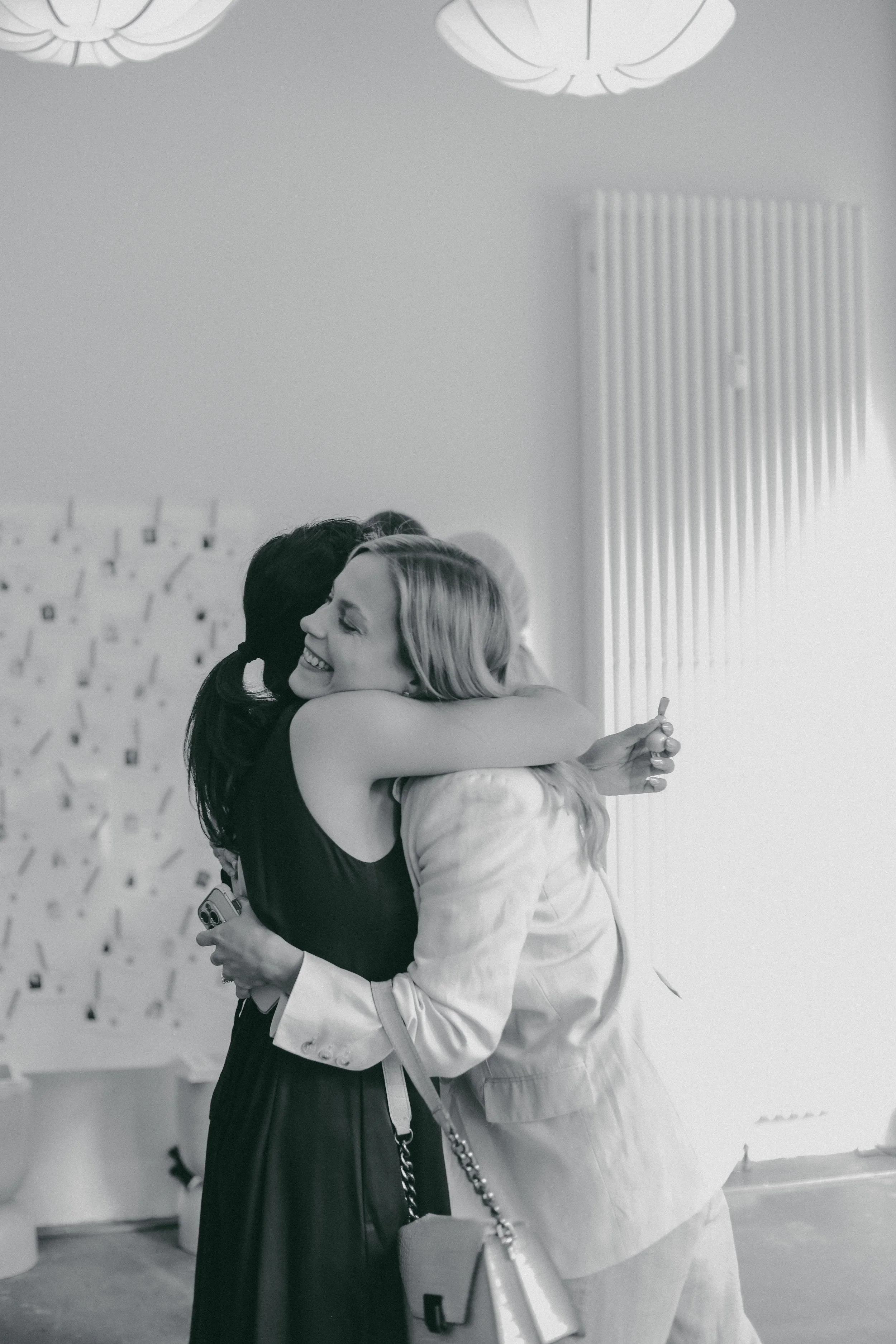 Zwei Frauen umarmen sich herzlich bei einer Begrüßung in einem Raum mit moderner Dekoration. Die Frauen lachen und scheinen glücklich.
