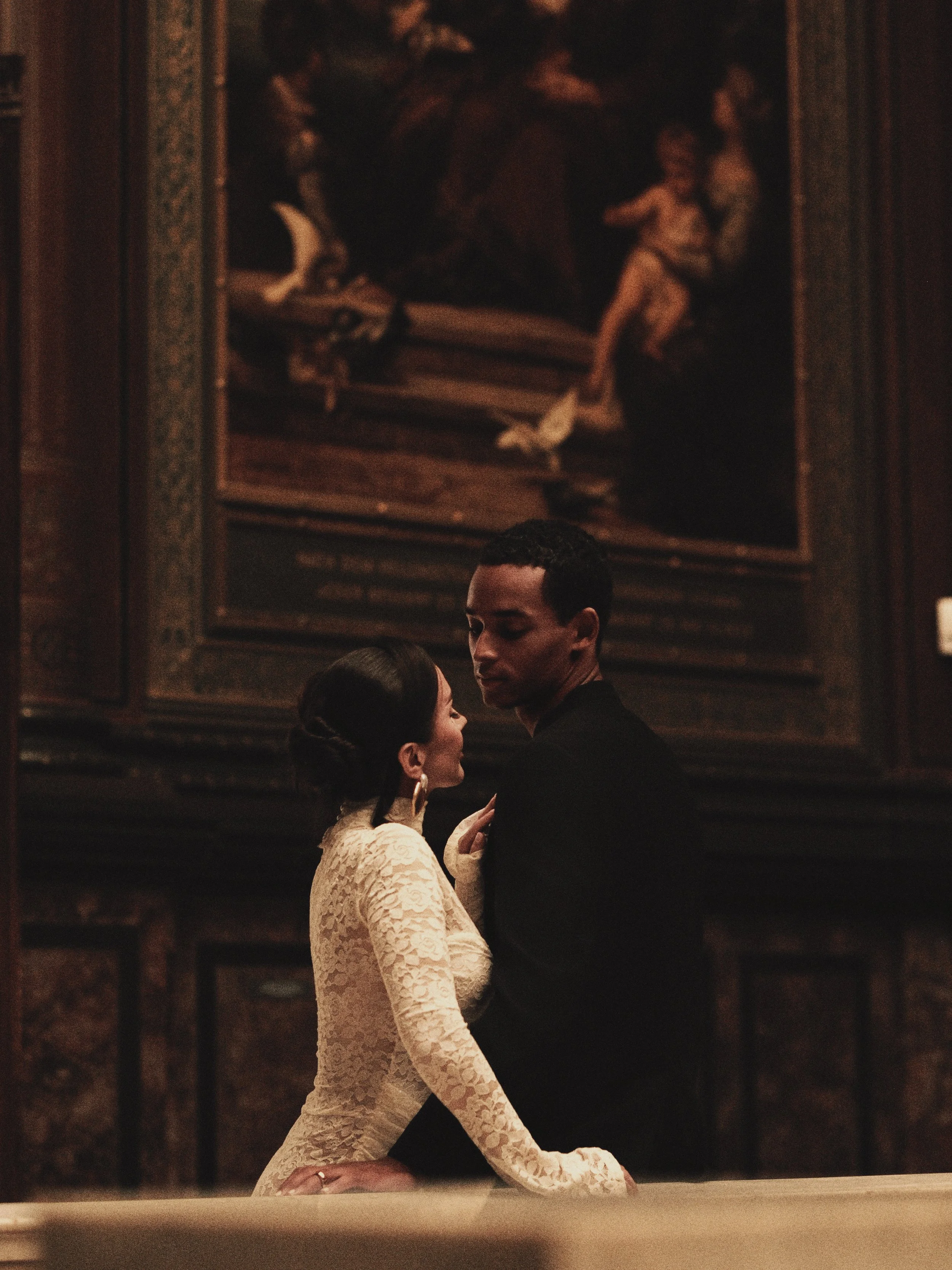 A woman in a lace dress and earrings and a man in a black outfit share an intimate moment in an art gallery or museum. They are positioned in front of a dark, richly decorated wall with a large, classical painting.