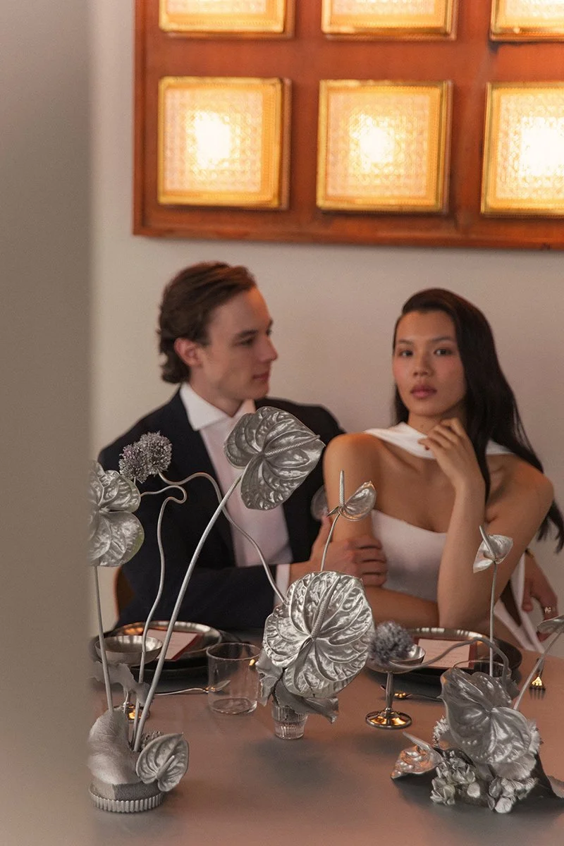 Ein elegantes Paar sitzt an einem festlich gedeckten Tisch in einem Restaurant, umgeben von silberfarbenen Dekorationen, im Hintergrund eine Wand mit goldumrandeten Bildern.