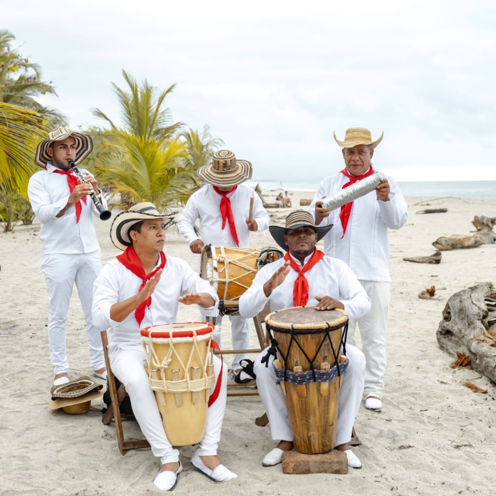 Grupo de músicos folclóricos tocando instrumentos en la playa, con sombreros, camisetas blancas y pañuelos rojos.