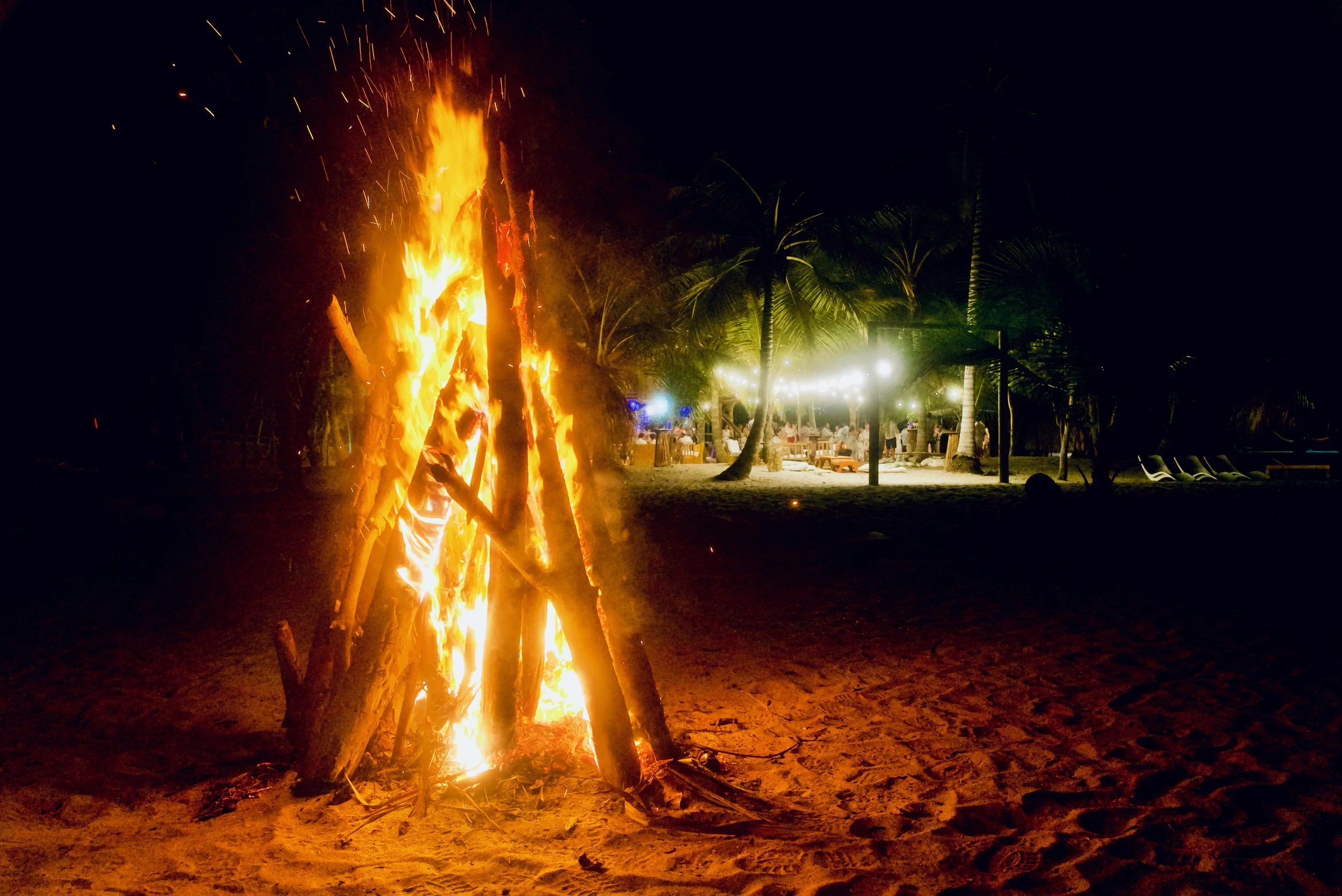 Fogata encendida en la playa nocturna con fondo de palmeras y un área con luces y mesas en la noche.