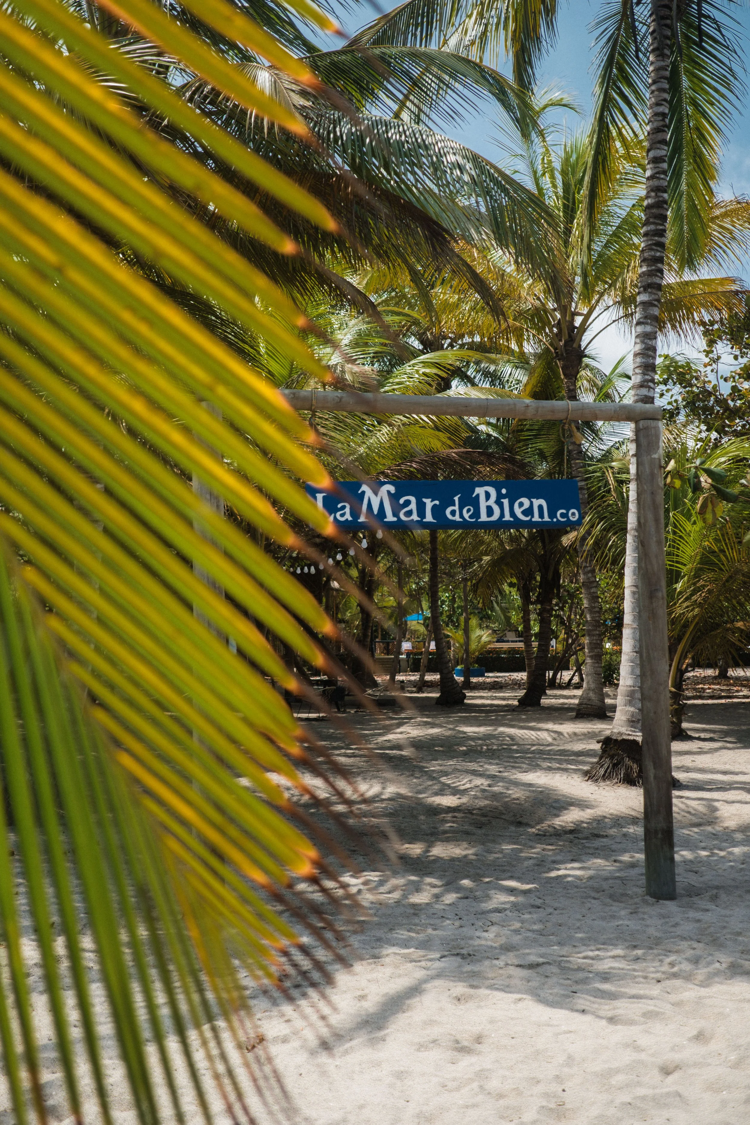 Cartel azul de bienvenida con letras blancas que dice "La Mar de Bien" en una playa privada con palmeras y arena.