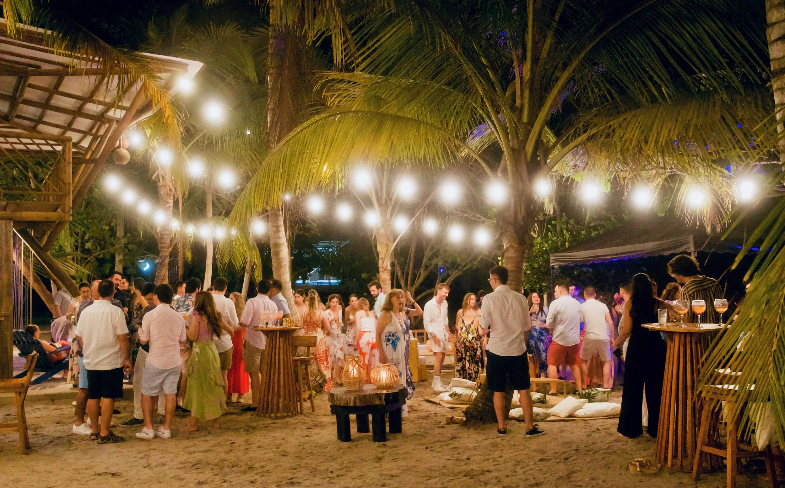 Personas en una fiesta al aire libre en la playa con lámparas colgantes y palmeras. preboda, postboda