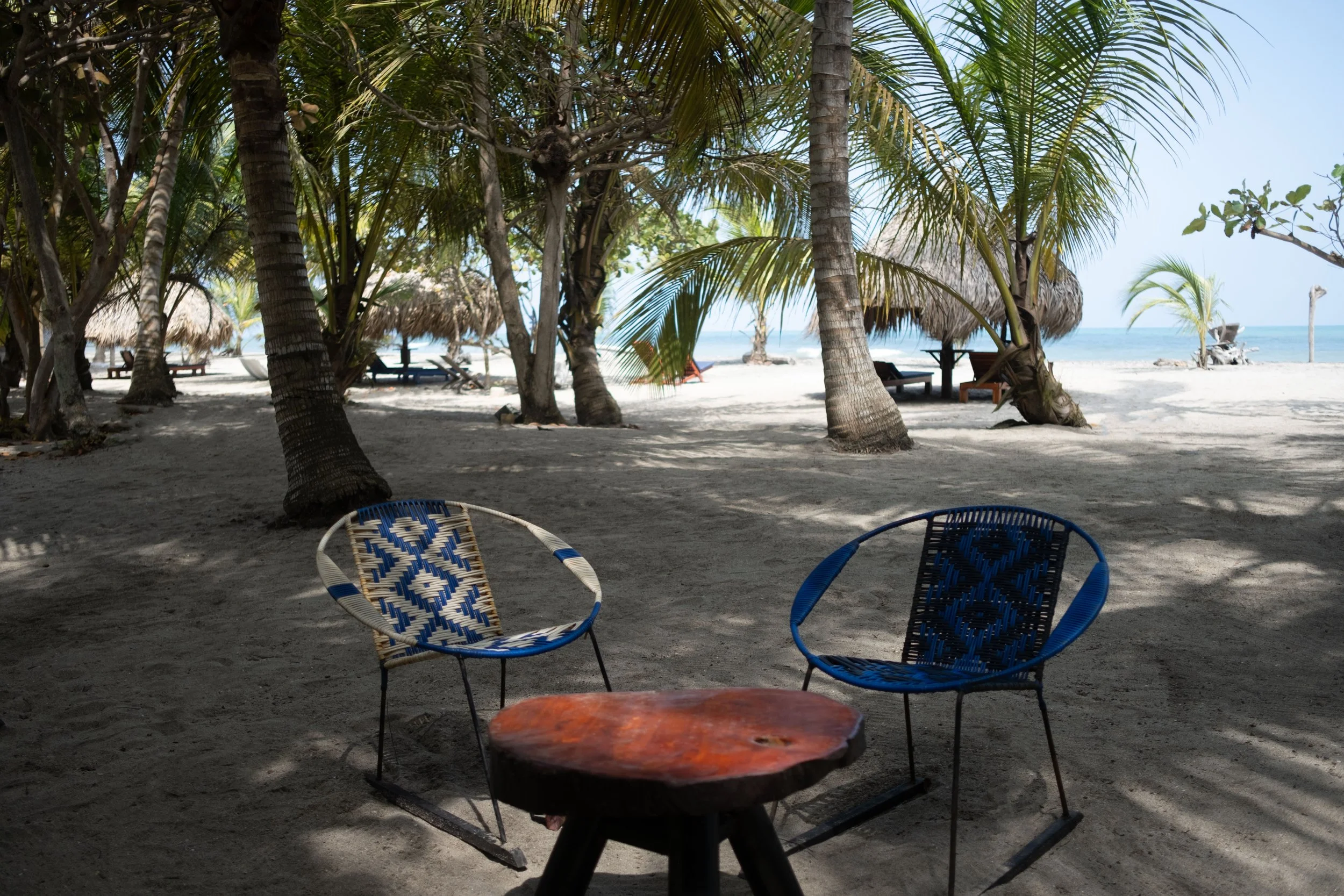 Dos sillas de mimbre en un área de playa con sombra de palmeras y vista al mar Caribe.