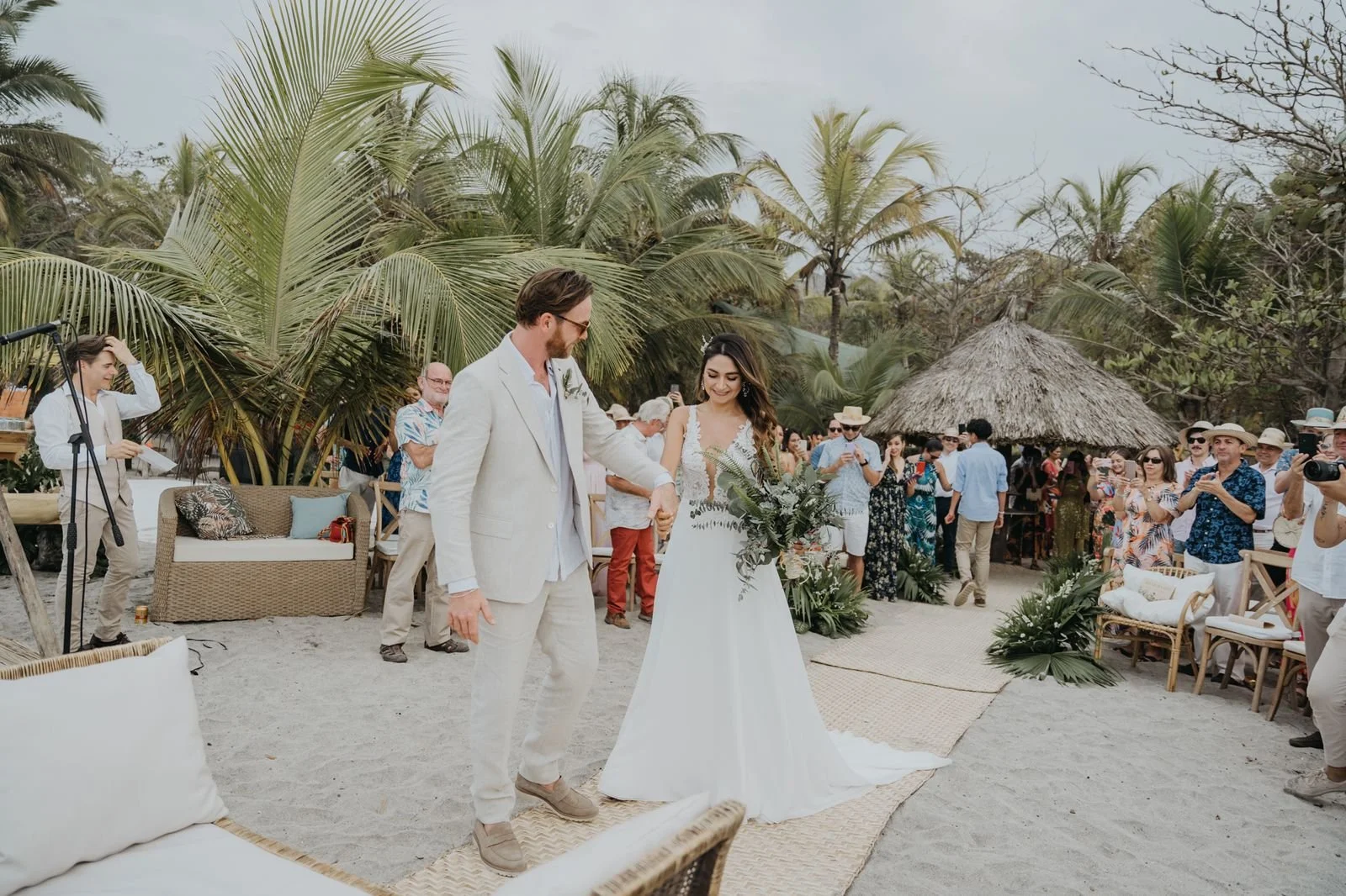 Una pareja se casando en una ceremonia en la playa rodeada de invitados y palmeras, en un entorno tropical.