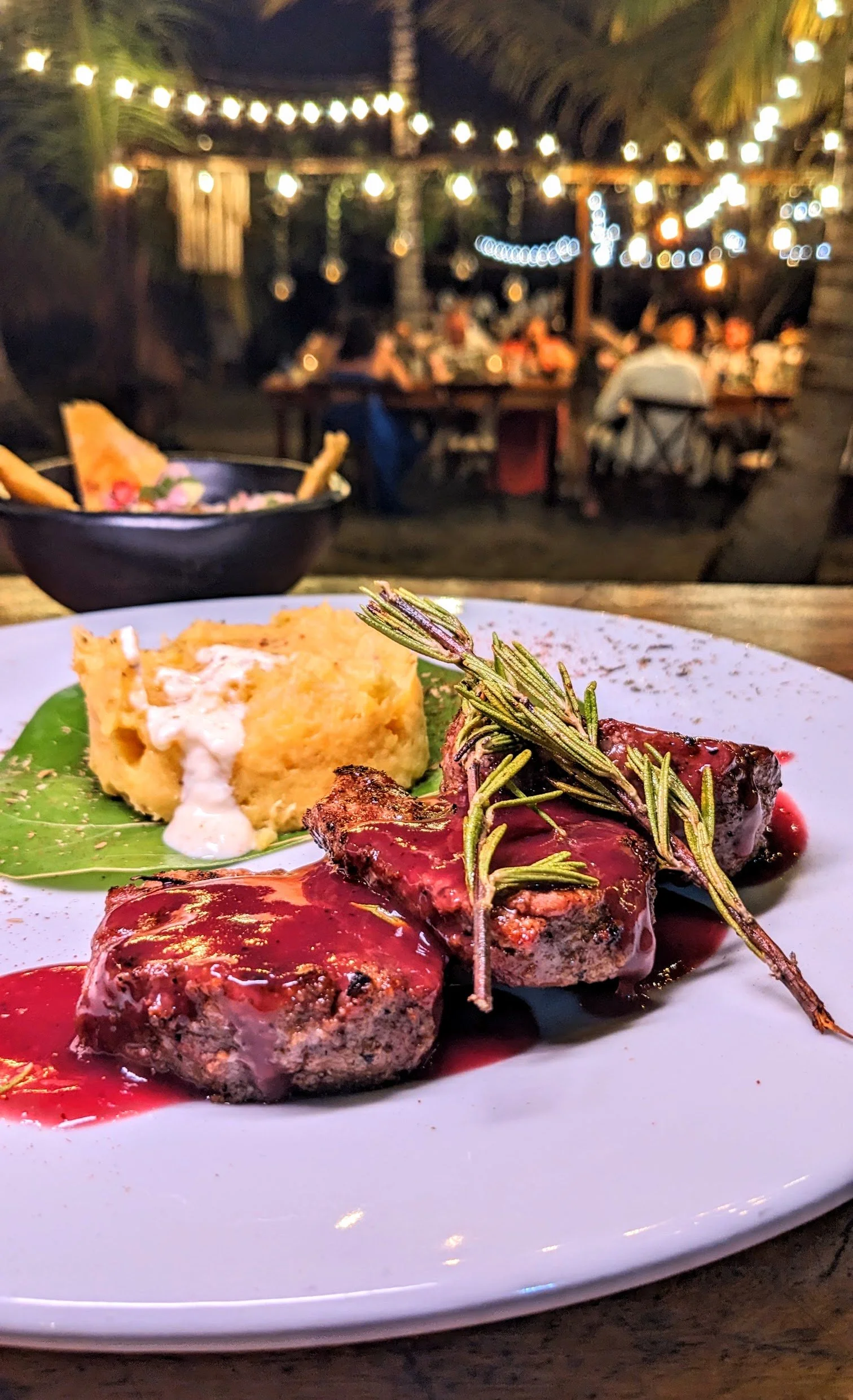 Plato de carne con salsa, puré de papas y una ramita de romero en un restaurante al aire libre de noche, con luces y comensales en el fondo.