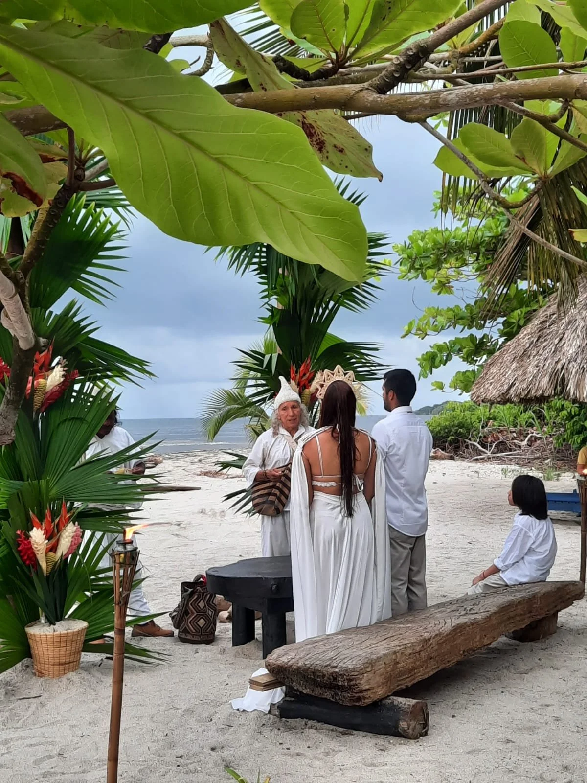 Ceremonia de matrimonio en la playa con personas vestidas de blanco, decoraciones tropicales y un fondo de mar y cielo nublado.