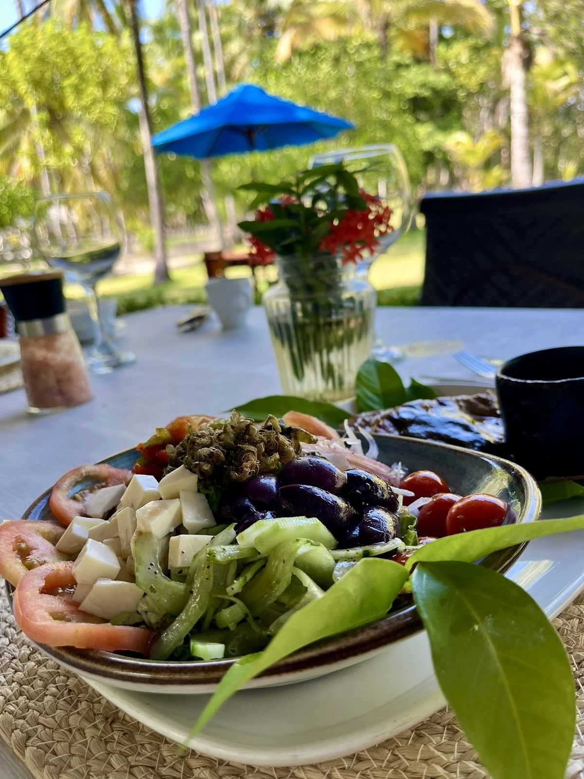 Plato gourmet con ensalada que incluye tomates, aceitunas negras, queso y verduras, en una mesa al aire libre con un jarrón de flores y un paraguas azul en el fondo.