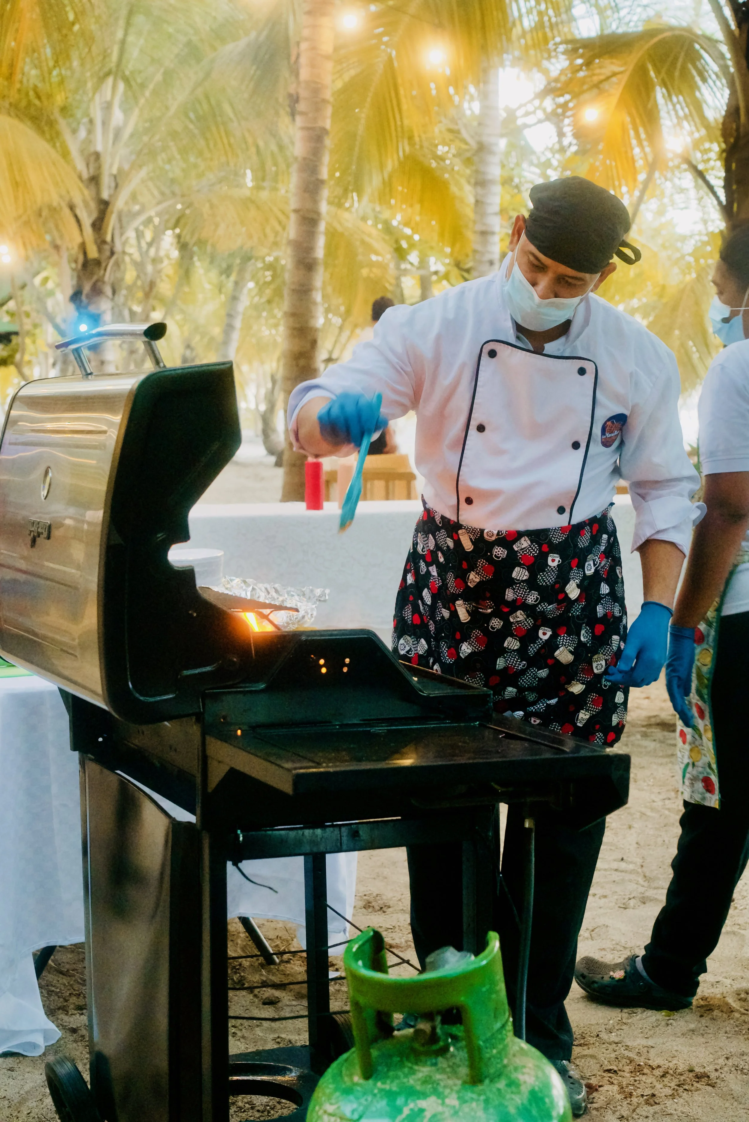 Chef cocinando en una parrilla al aire libre con fondo de palmeras.