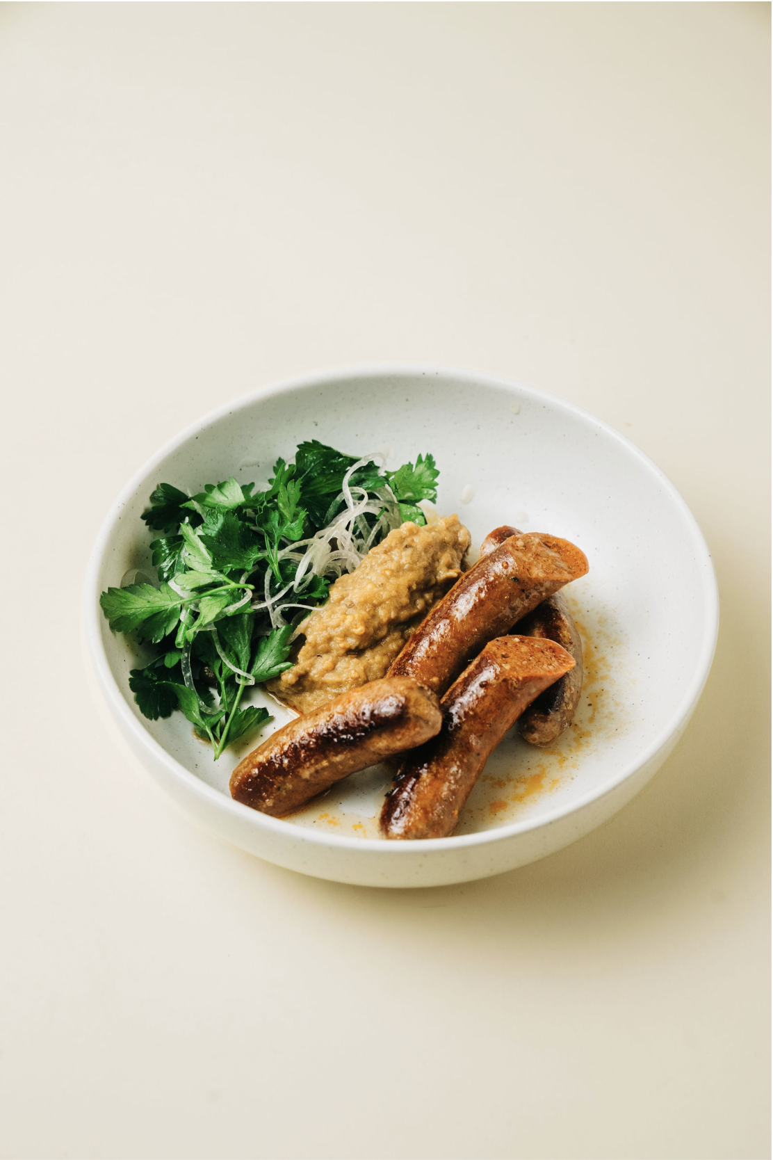 A white bowl containing grilled sausages, a dollop of mustard or horseradish, and fresh herbs including parsley and cilantro.