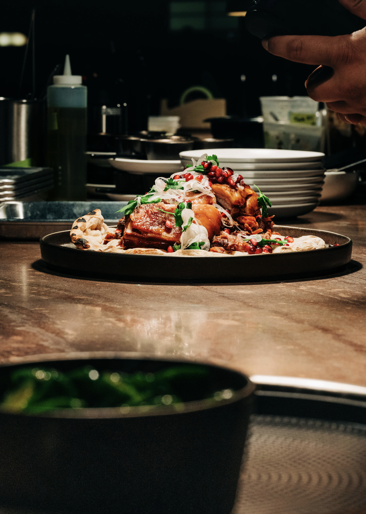 A plated gourmet dish topped with pomegranate seeds, herbs, and sliced onions on a dark plate, with a chef working in the background in a professional kitchen.