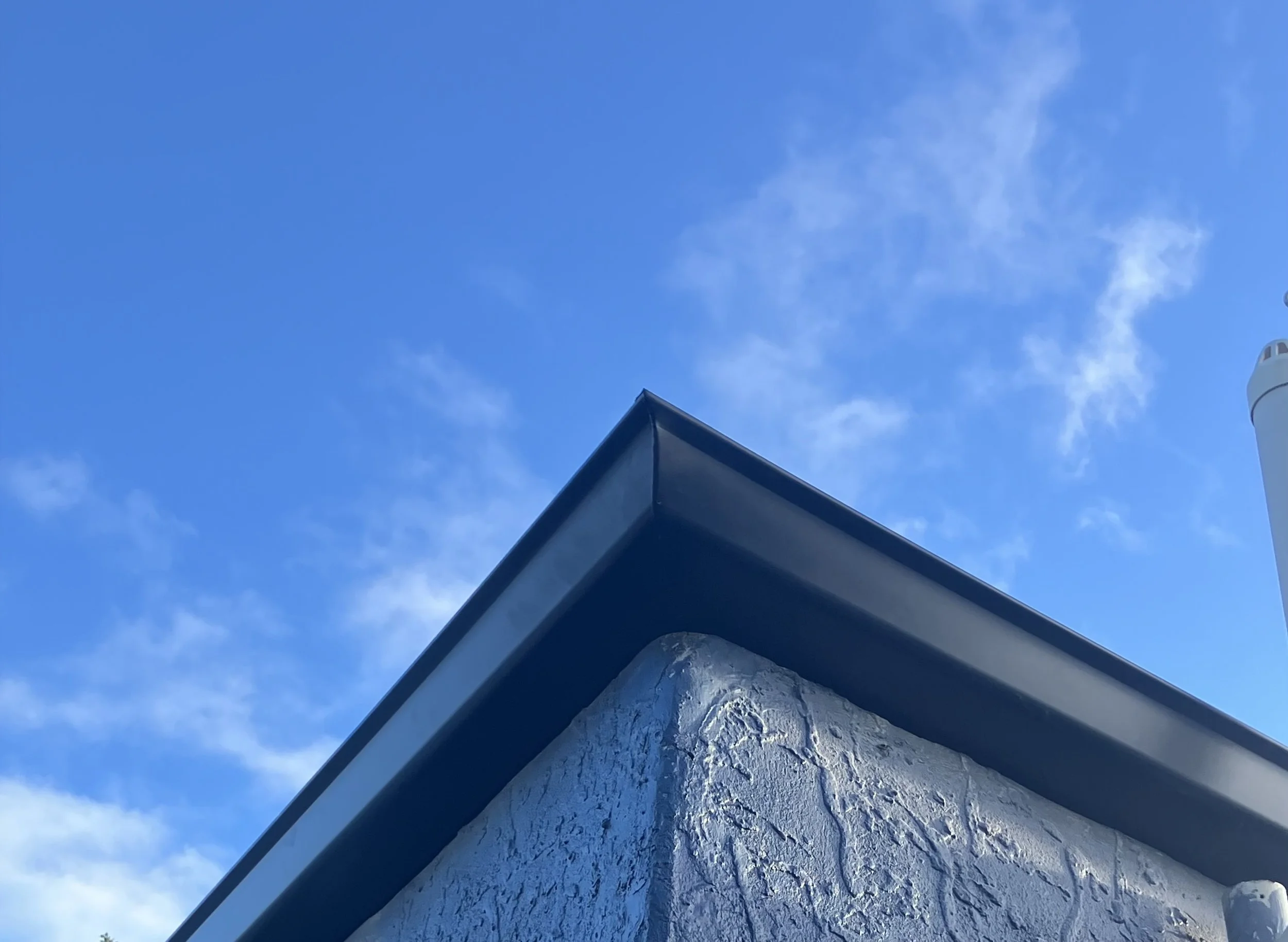 Close-up of a building corner with textured white stucco wall and metal roof edge against a background of a blue sky with wispy clouds.