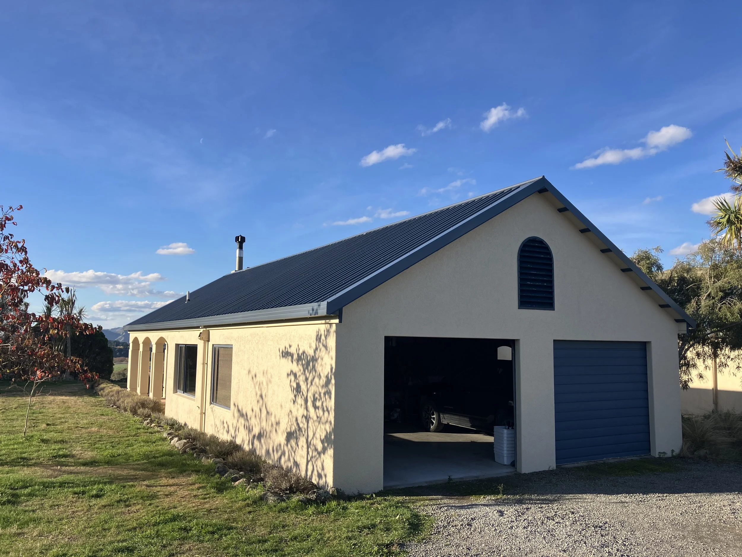 A beige building with a blue metal roof, garage door, and small arched window, set against a clear blue sky with a few clouds, surrounded by grass and trees.