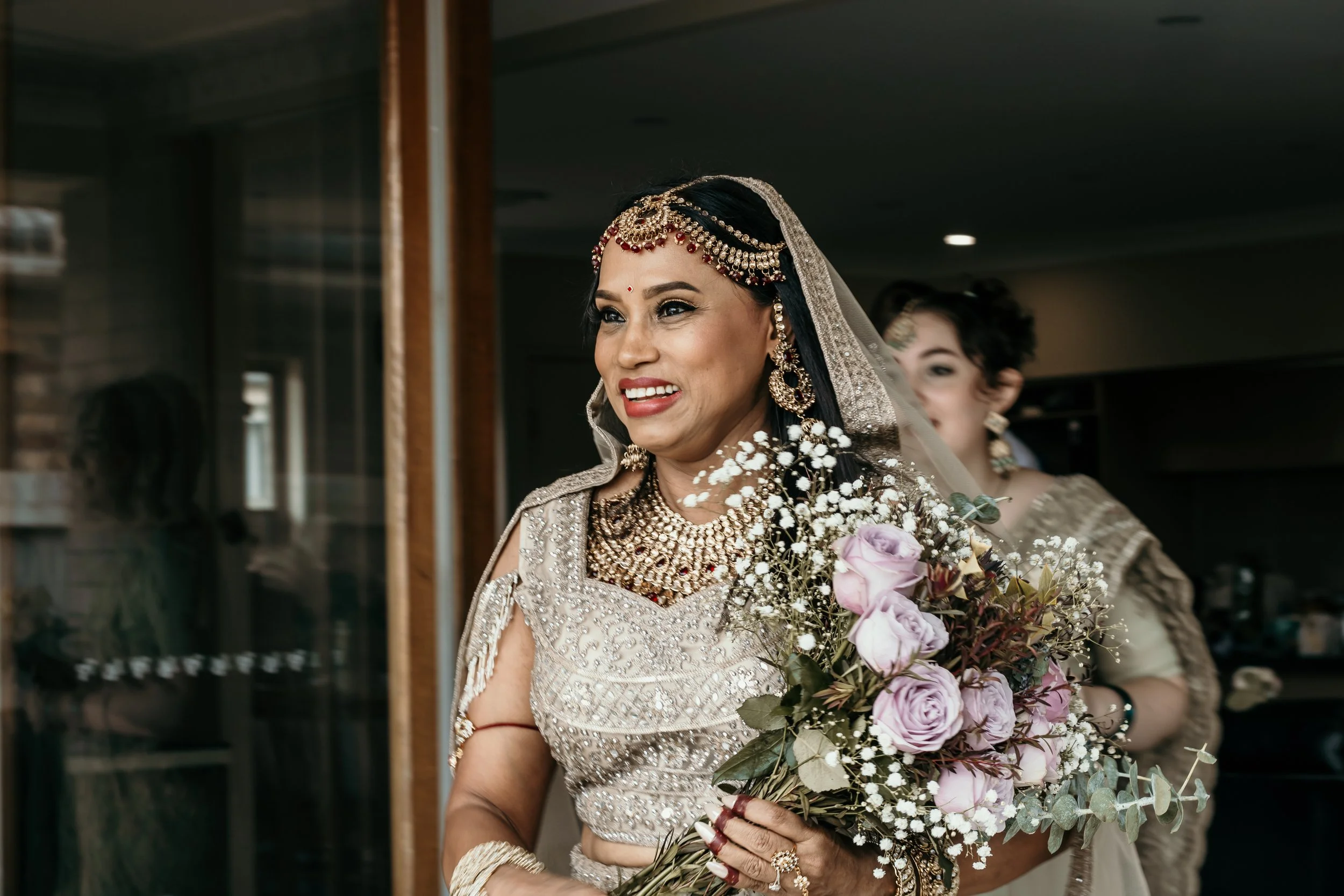 A woman dressed in traditional Indian bridal attire holding a bouquet of light purple roses, with another woman in similar attire visible in the background.