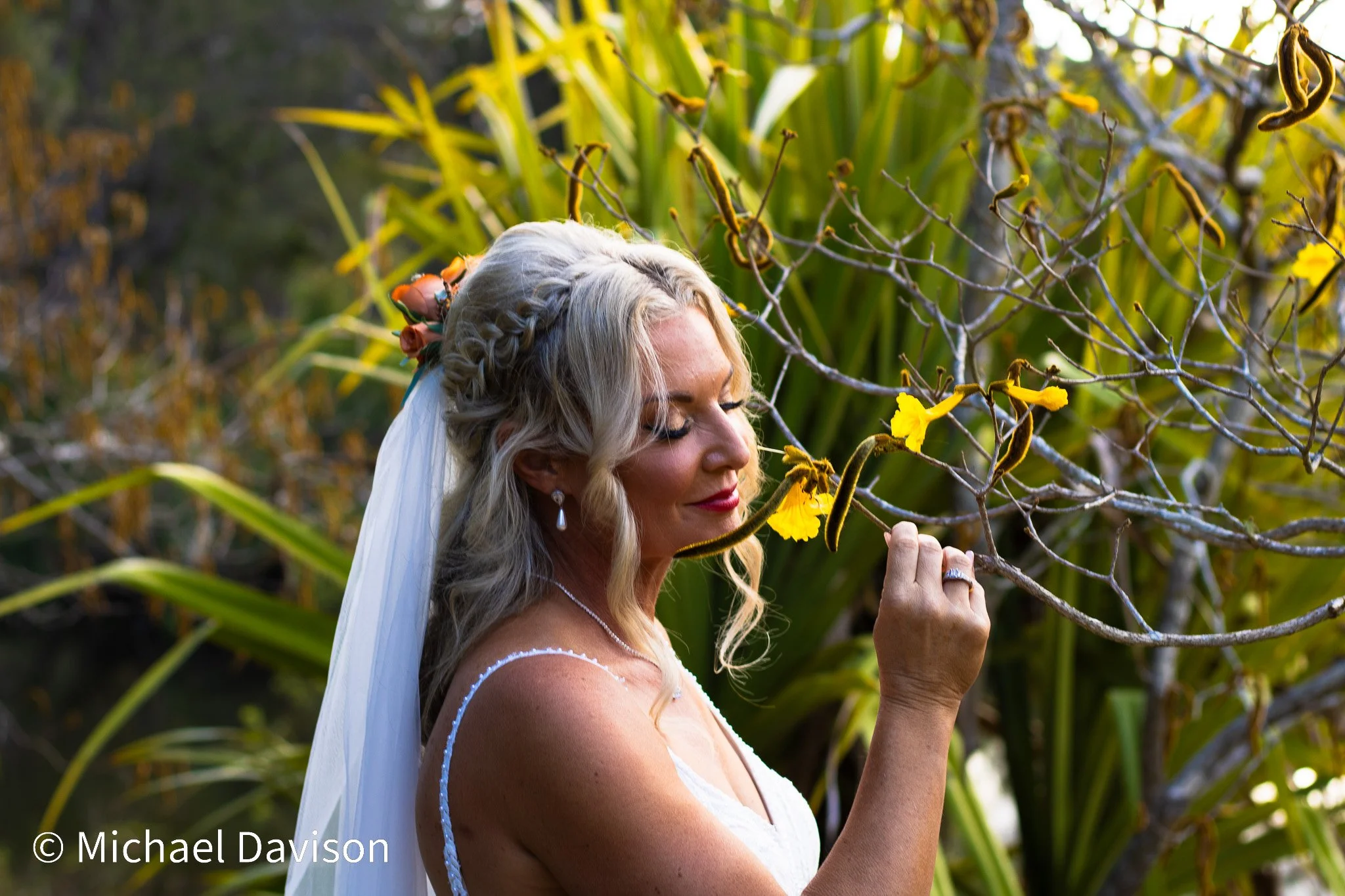 A woman with long blonde hair, wearing a white dress and veil, holds a yellow flower on a tree branch and touches it with her face, surrounded by greenery and plants.