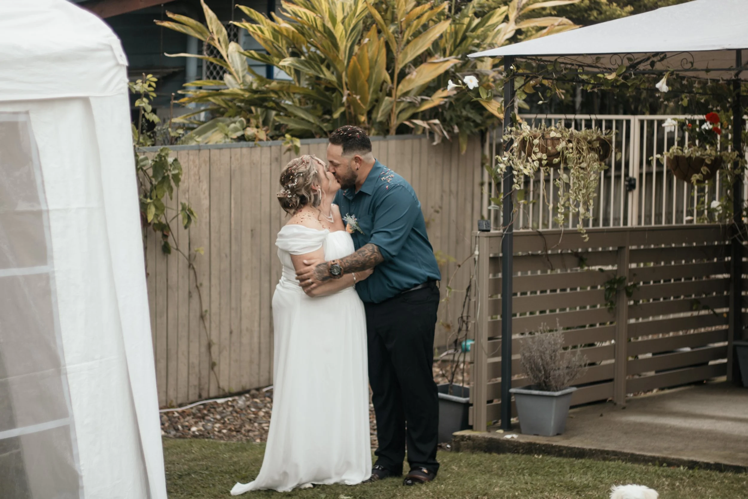 A couple is kissing outdoors in a garden or backyard, dressed in wedding attire. The woman is in a white dress, and the man is in a blue shirt and black pants.
