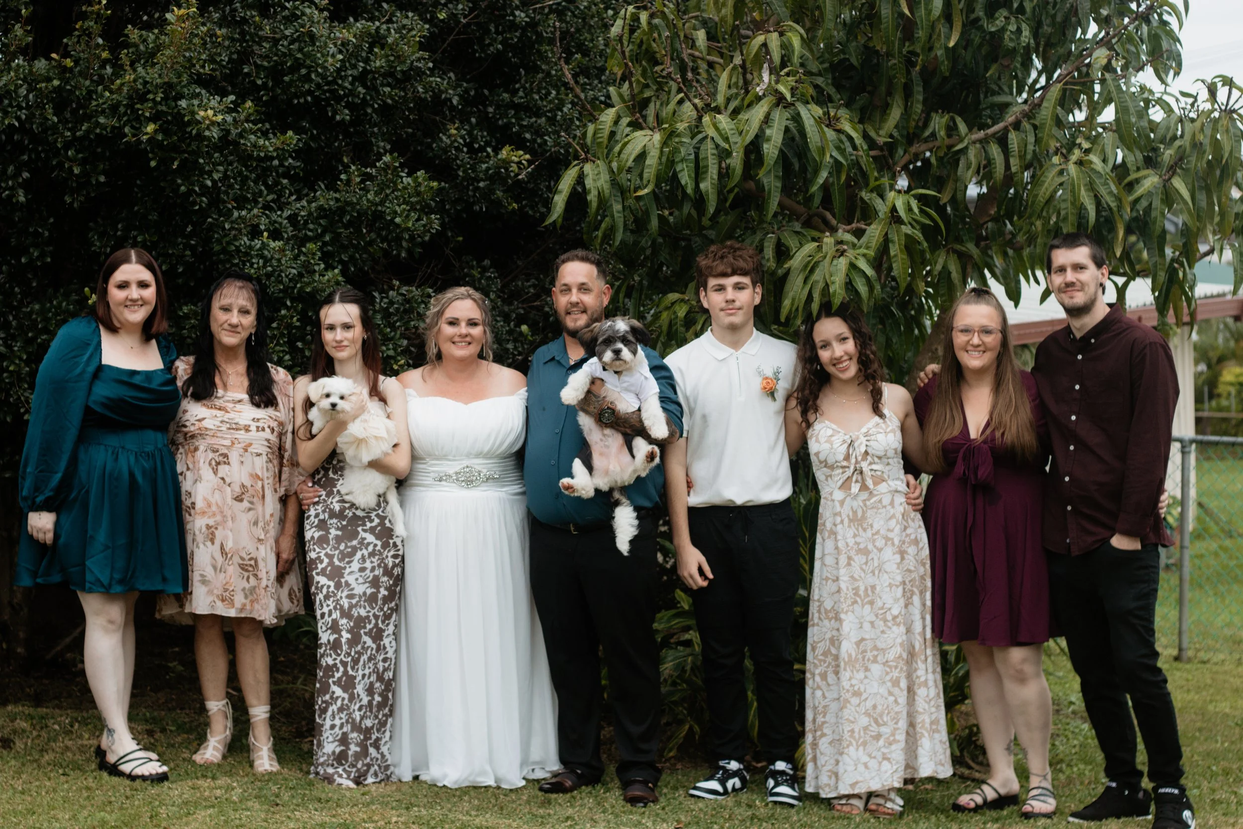 Group of eleven people and two dogs standing outdoors in front of a tree, dressed in semi-formal attire for a celebration or gathering.