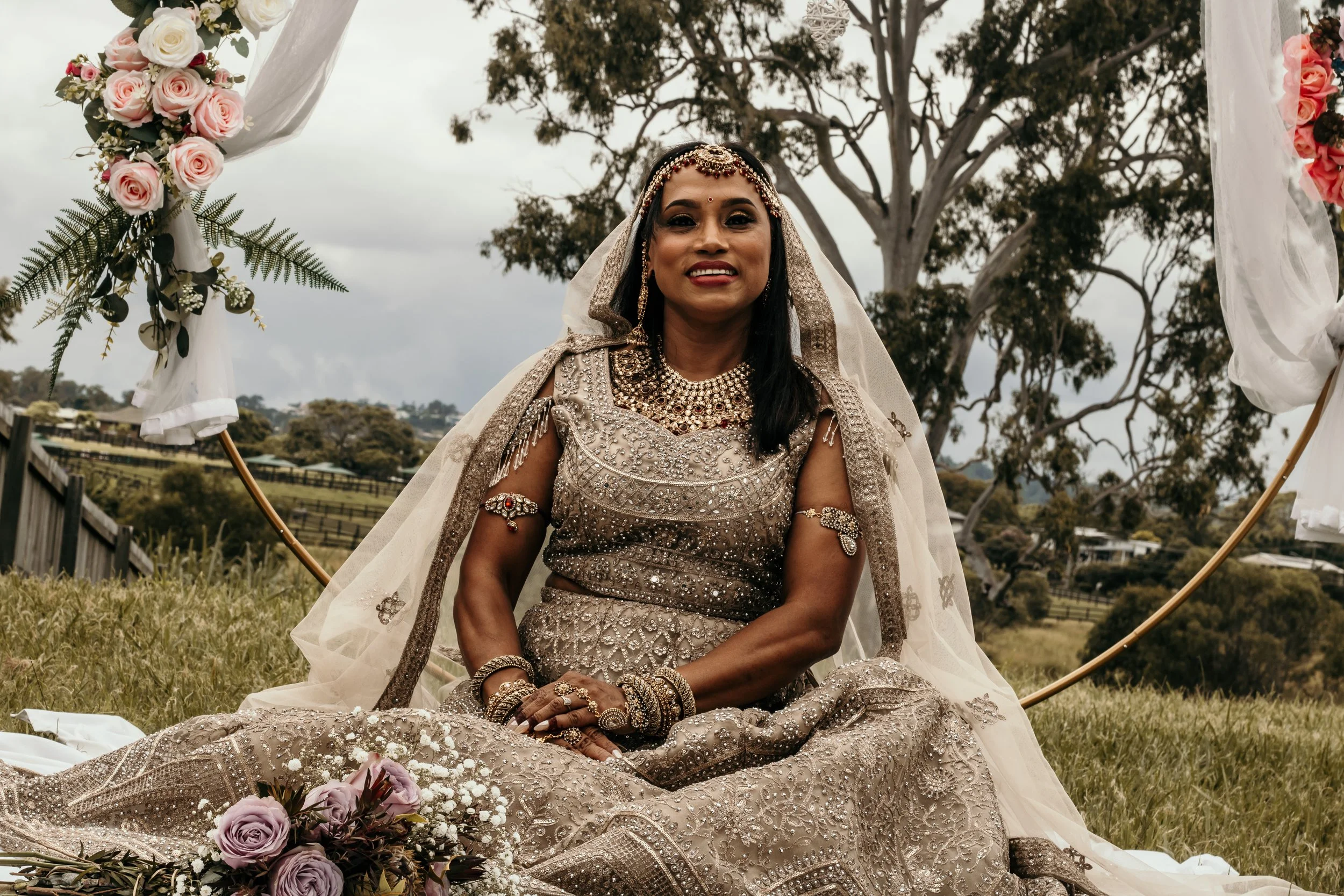 A woman dressed in a traditional, heavily embellished gown with jewelry sitting outdoors with trees and cloudy sky in the background. She is surrounded by floral decorations.