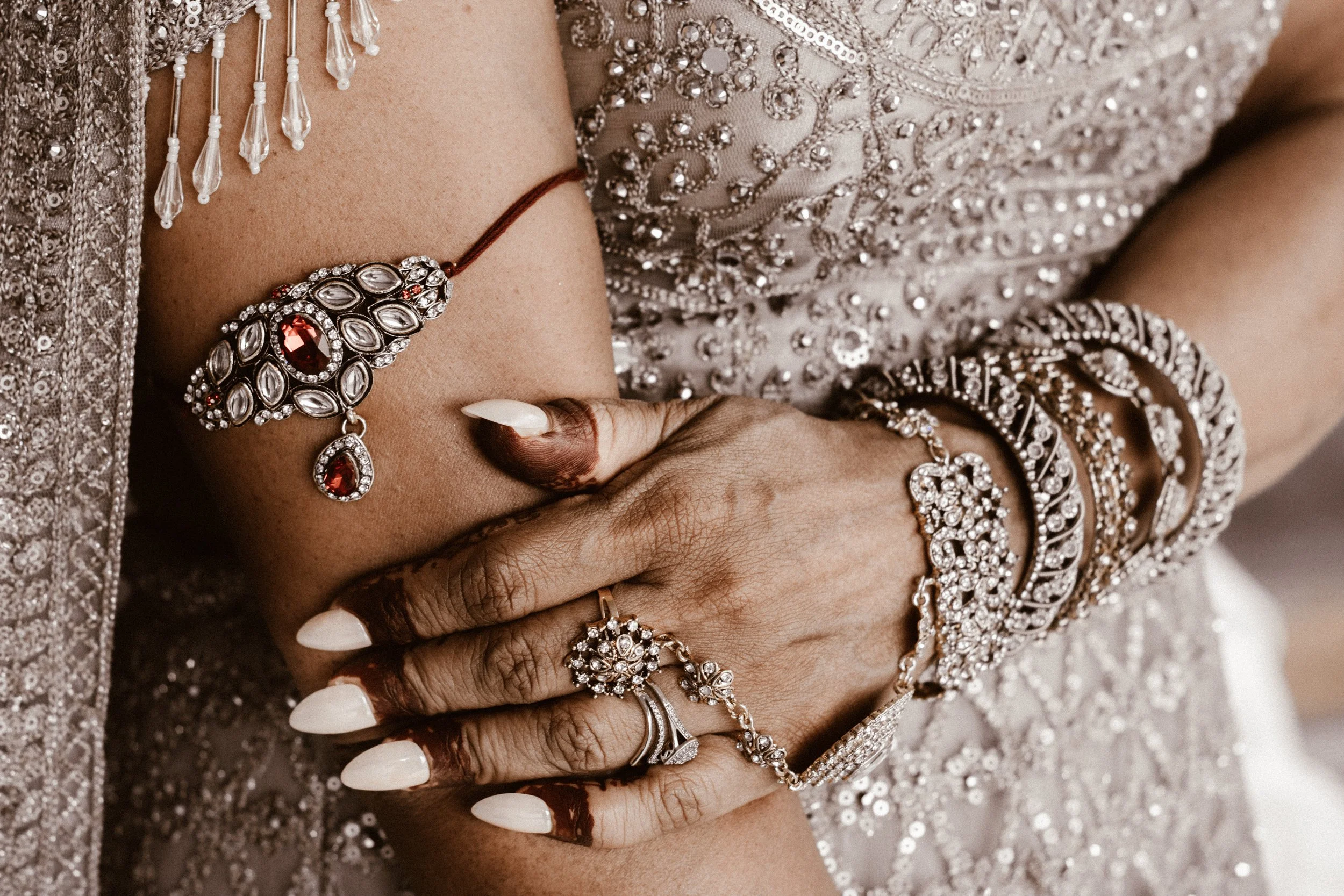 Close-up of a woman's hand and arm adorned with jeweled rings, bangles, and an ornate necklace, with a decorated traditional Indian attire in the background.