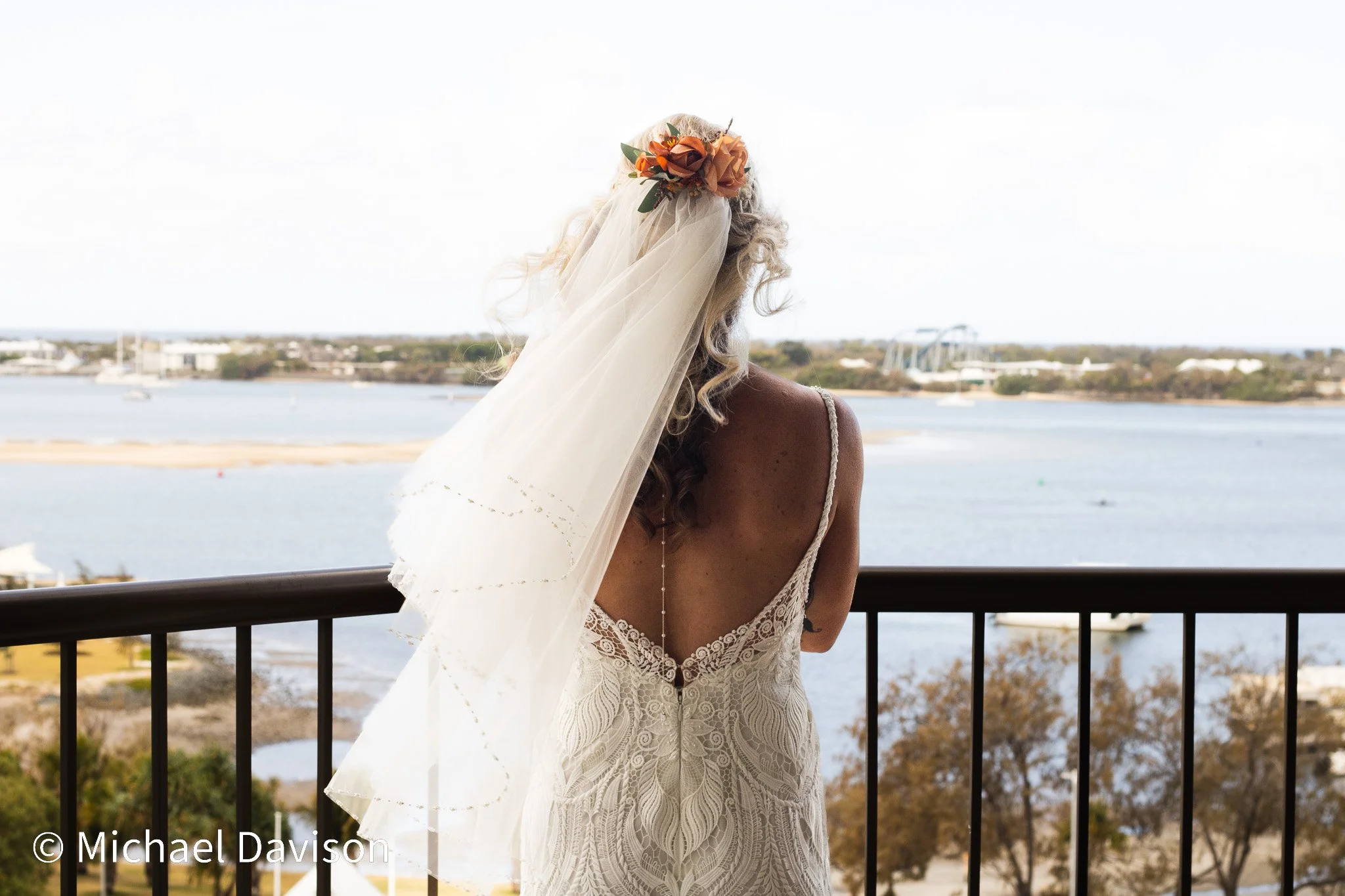 A woman wearing a wedding dress and veil, holding a floral headpiece, stands on a balcony overlooking a river or bay with boats and trees.