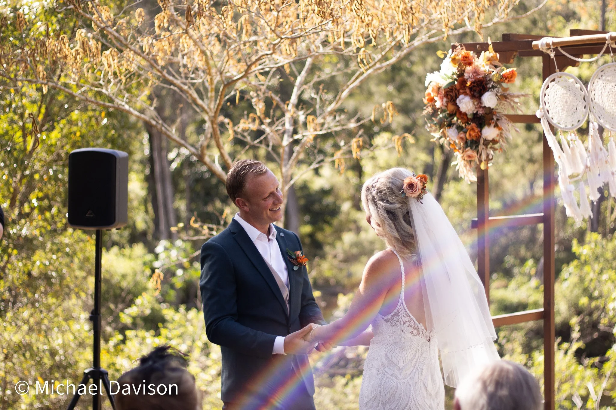 A bride and groom holding hands during their outdoor wedding ceremony. The groom is wearing a dark suit, and the bride is wearing a white lace dress with a veil and flower hair accessory. There is a floral arrangement and dreamcatchers on a wooden ar