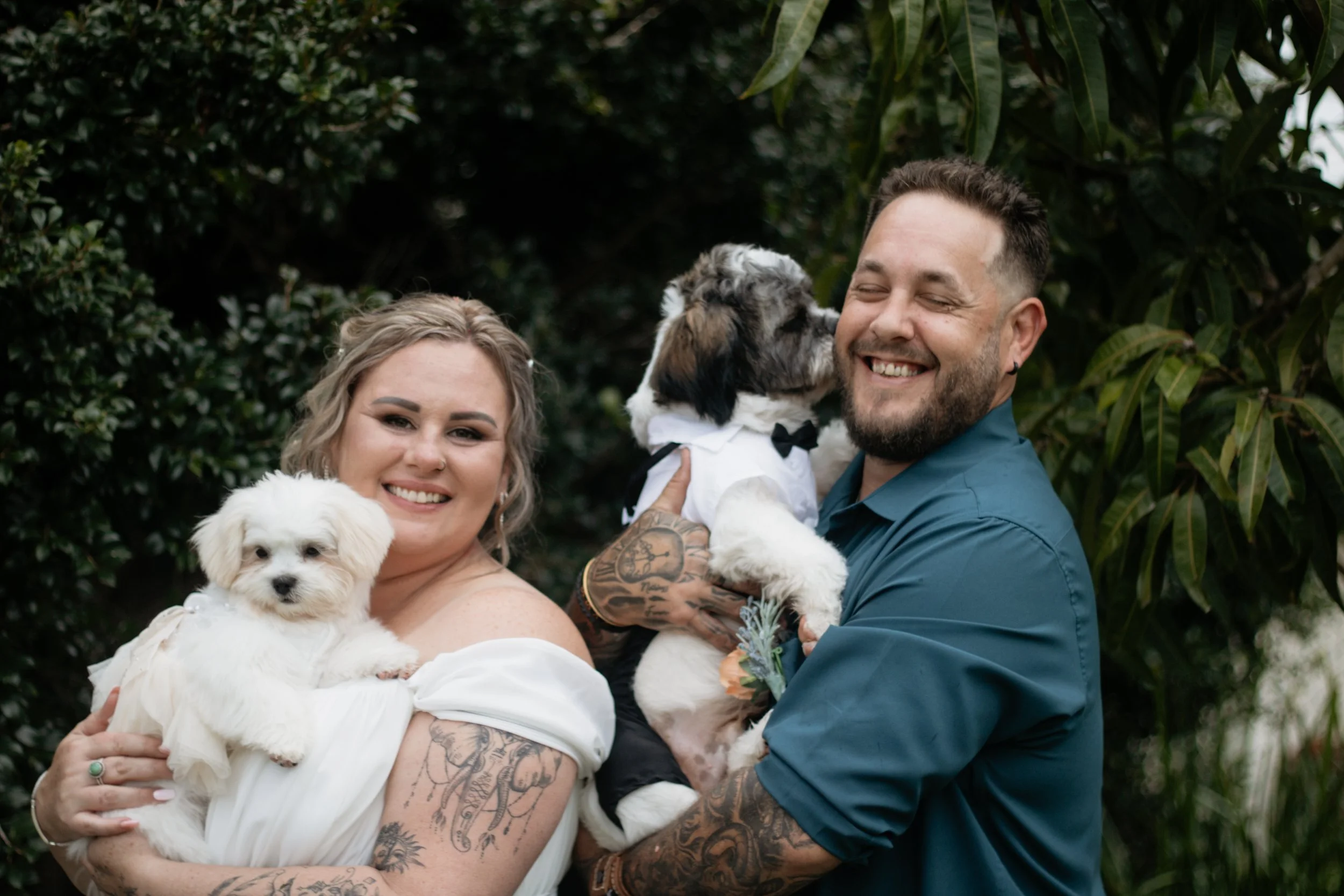 Smiling woman and man holding dogs outdoors, surrounded by green foliage.