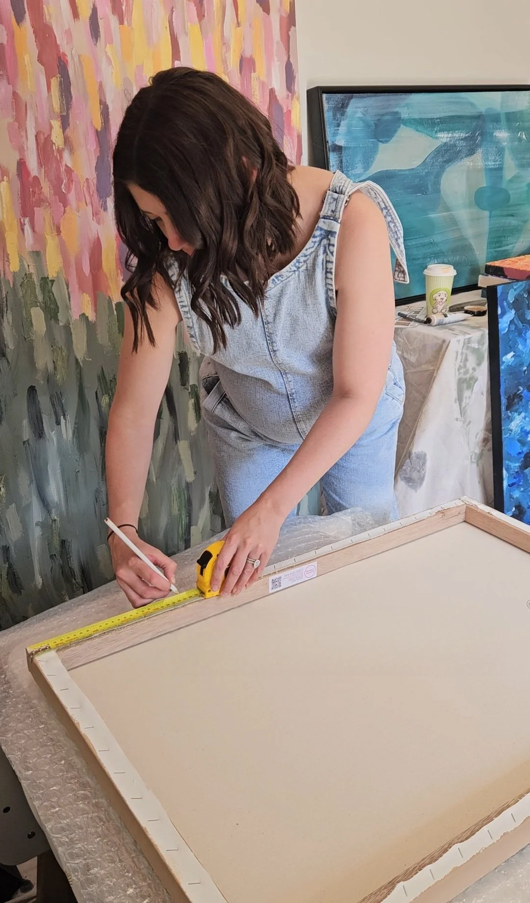 A woman with dark brown hair measuring and marking a wooden frame on a table in front of her, with abstract paintings and art supplies in the background.