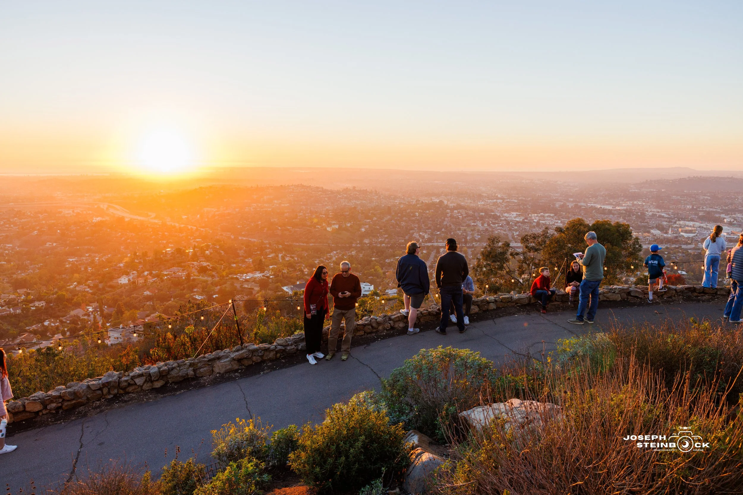 People gathered on a lookout at sunset, overlooking a cityscape from a hill with plants and rocks in the foreground.