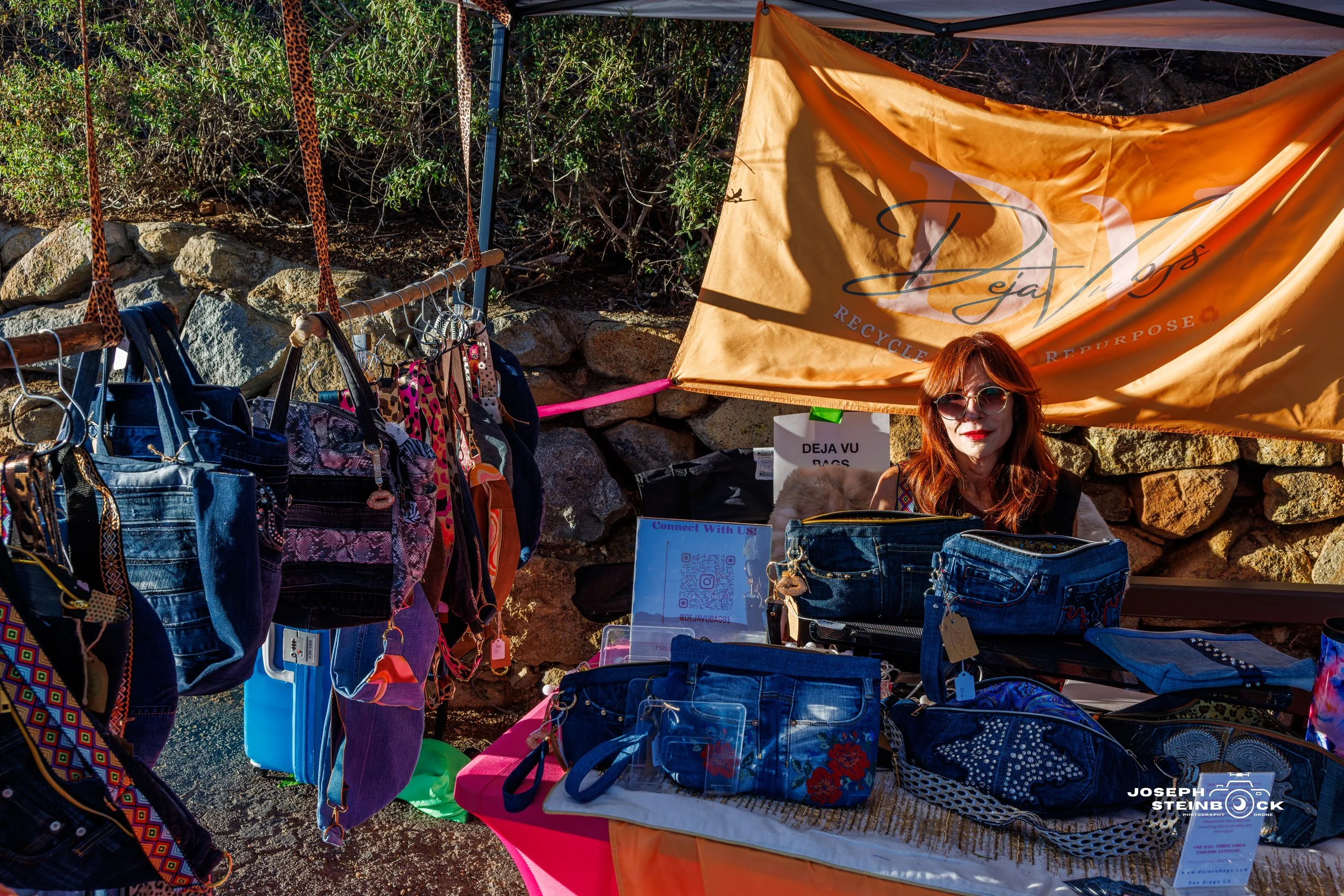 A woman with red hair and sunglasses sitting behind a booth of denim bags and accessories at an outdoor market. A tan banner with the logo and words 'Reclaim. Recycle. Repurpose.' is behind her. Various bags are displayed on the table and hanging on 