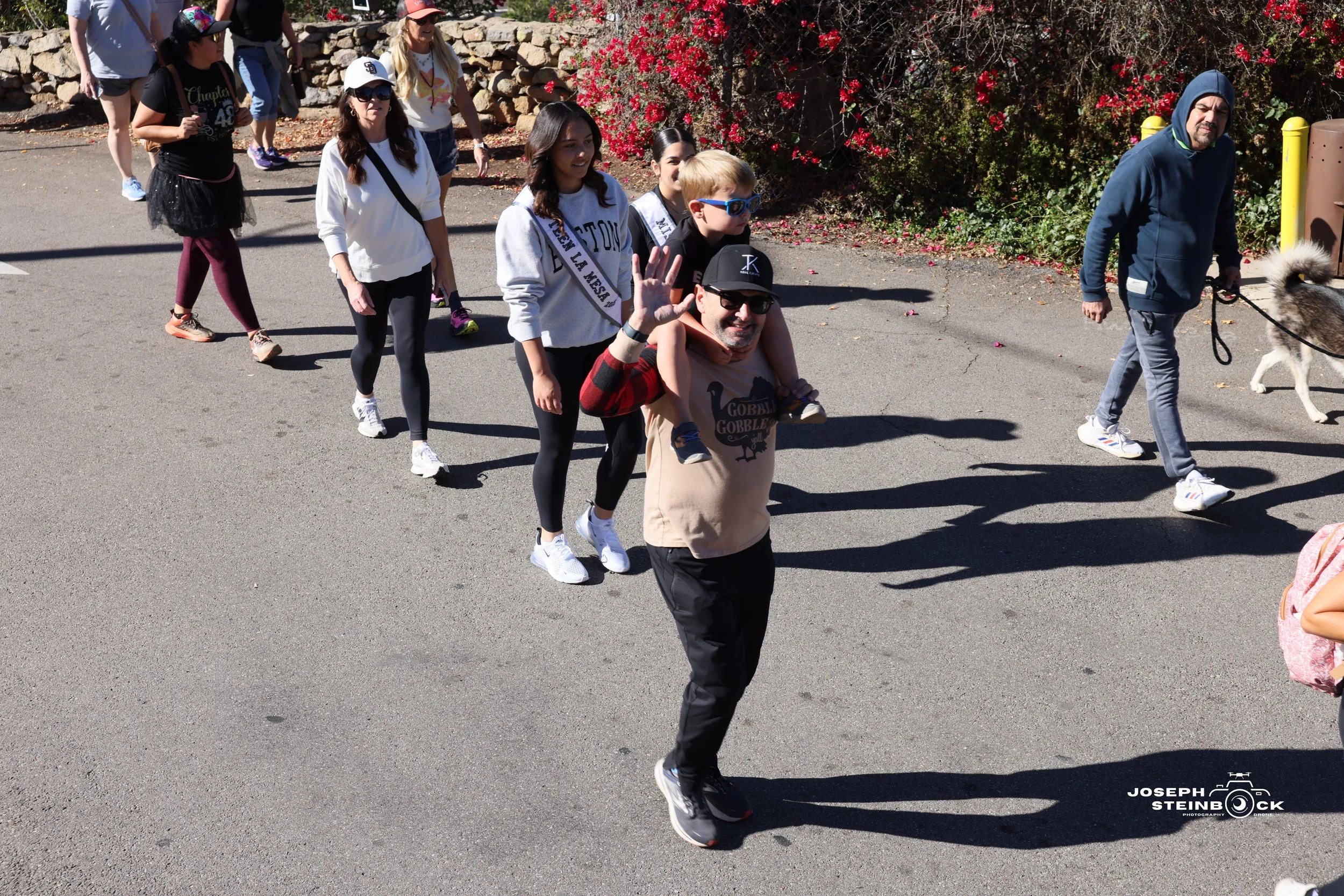A group of people walking outdoors on a sunny day, with some individuals wearing sunglasses and casual clothing. A man in the front is carrying a child on his shoulders, both smiling and waving at the camera. Other adults and children follow behind, 