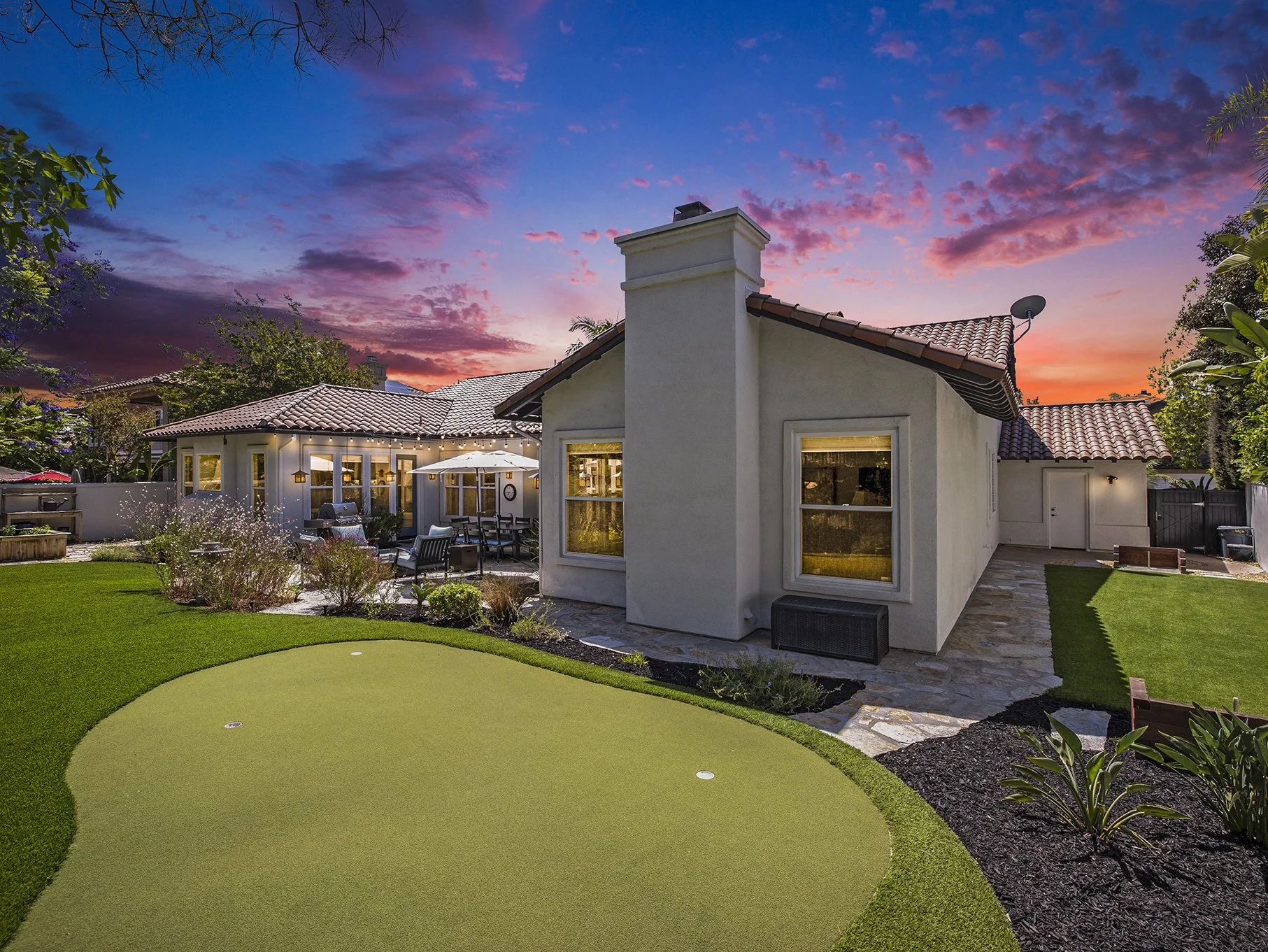 Backyard of a house with a putting green, patio furniture, and a sunset sky.
