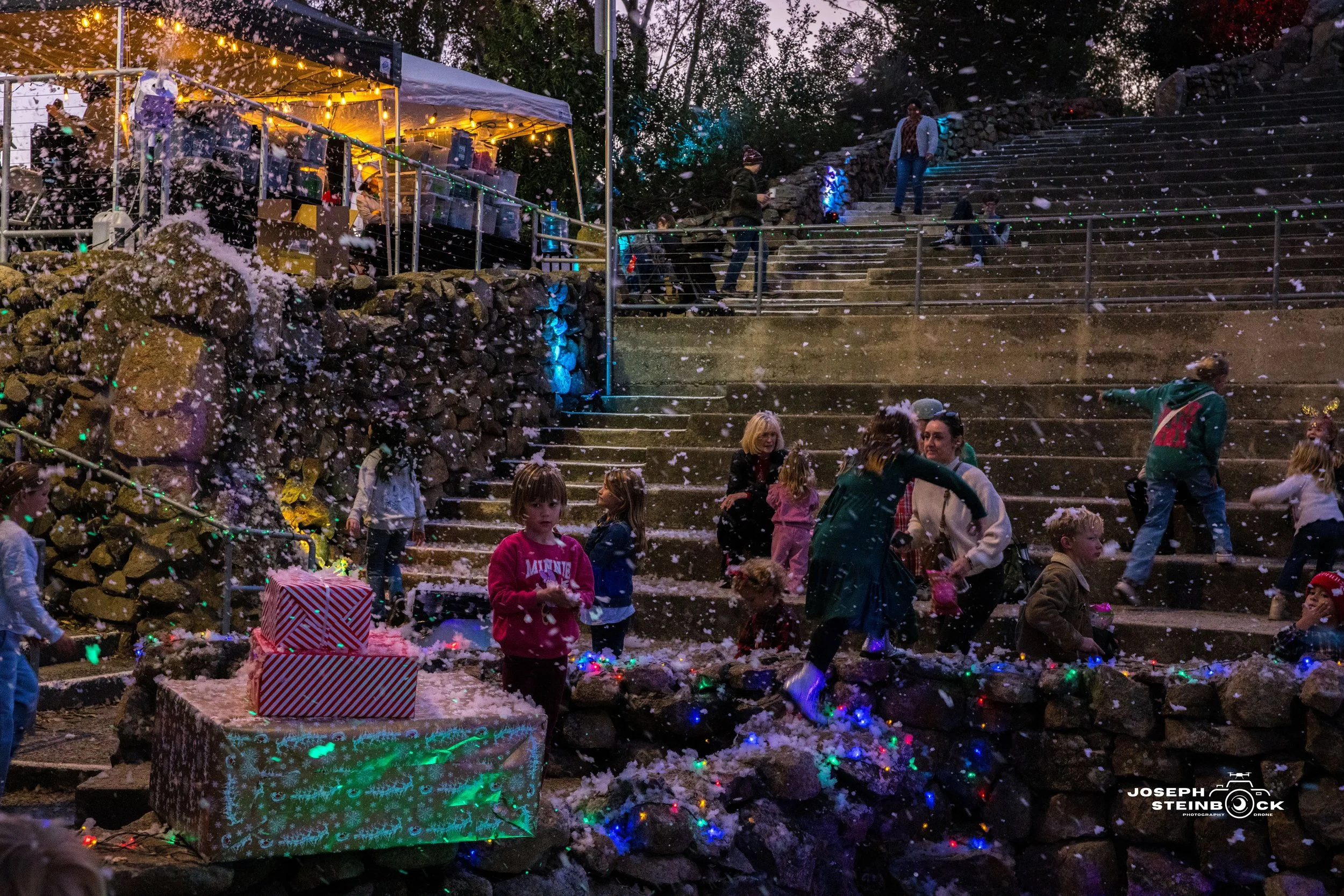 Children and adults celebrating outdoors during winter evening, with snow falling, Christmas decorations, and colorful string lights on a stone staircase and a tent with bright lights.