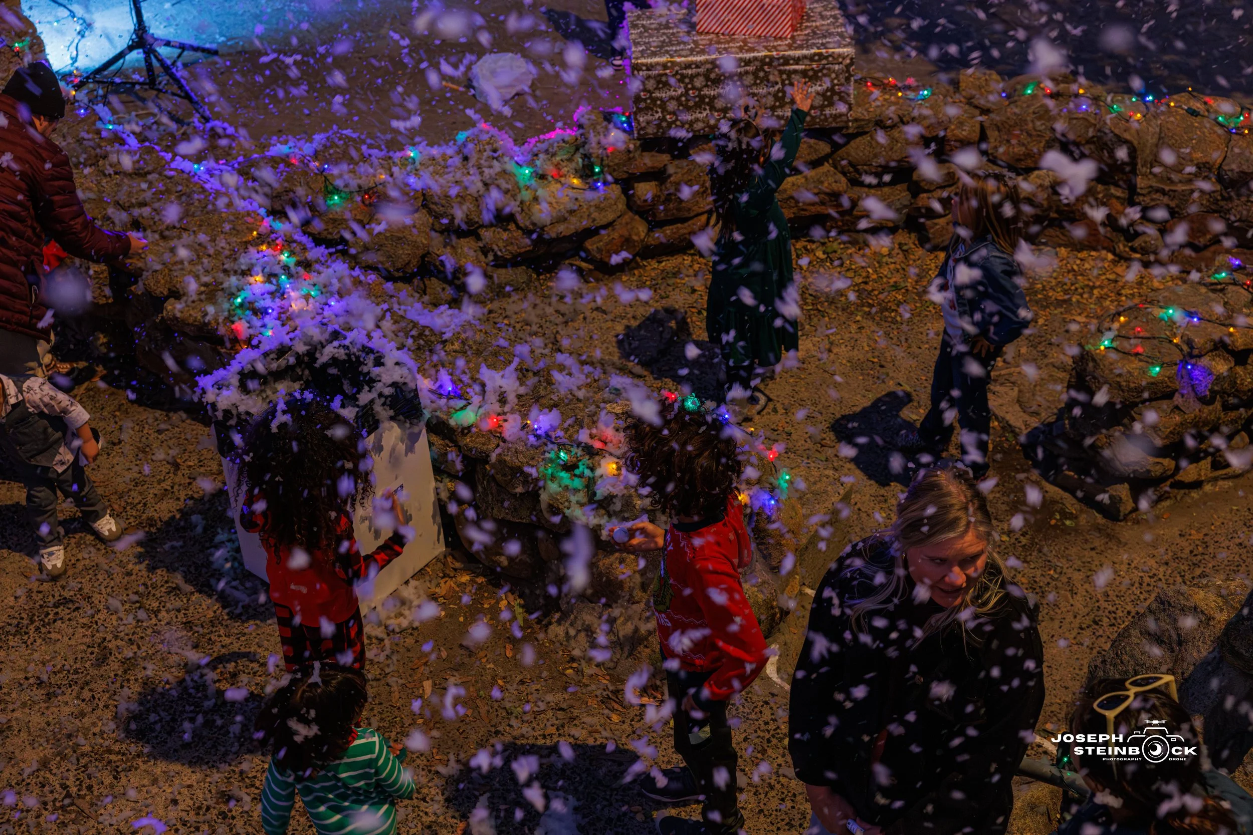 People celebrating outdoors at night with snow falling, colorful lights, and a stone wall