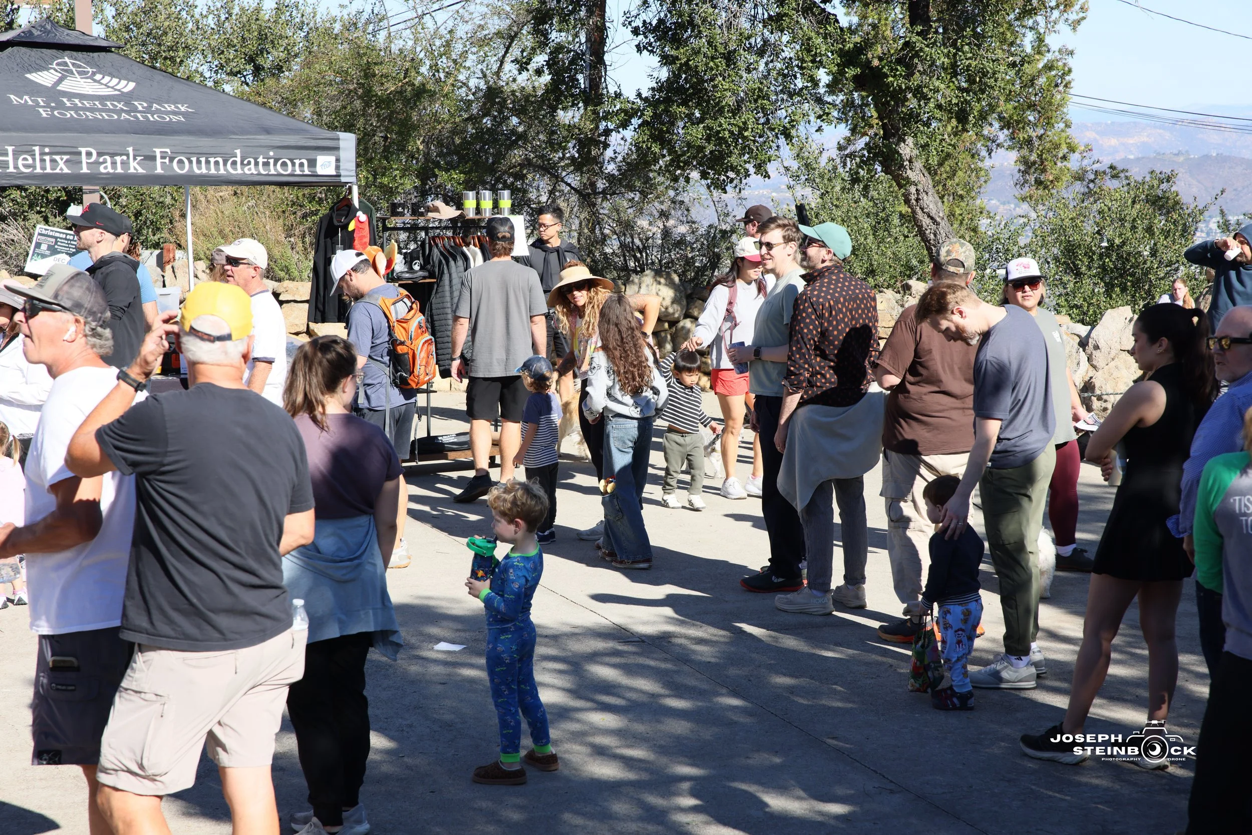 People gathered outdoors at a booth with a sign for the Helix Park Foundation, under a black canopy, with trees and mountains in the background.