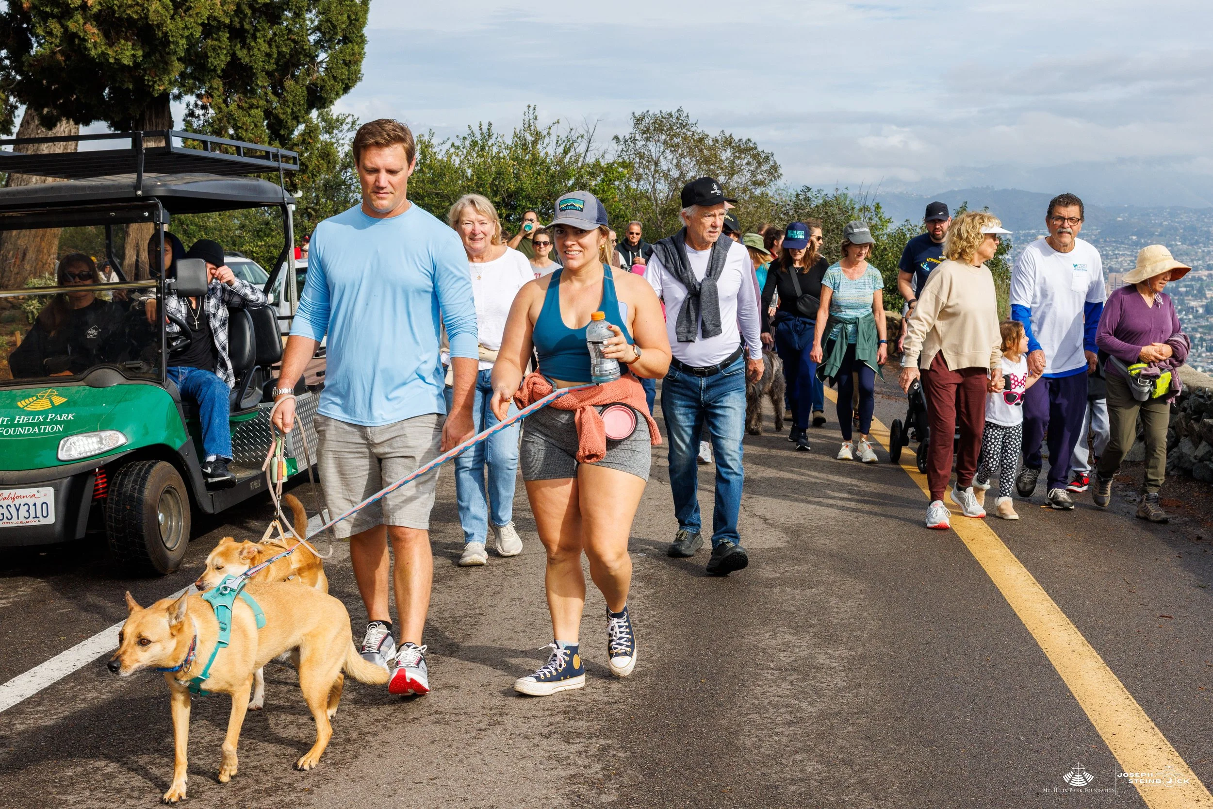 A group of people walking outdoors, some with dogs, on a paved path near trees and a distant view of a city or town. Some people are holding hands, wearing casual clothing, and the scene appears to be a community event or walk.