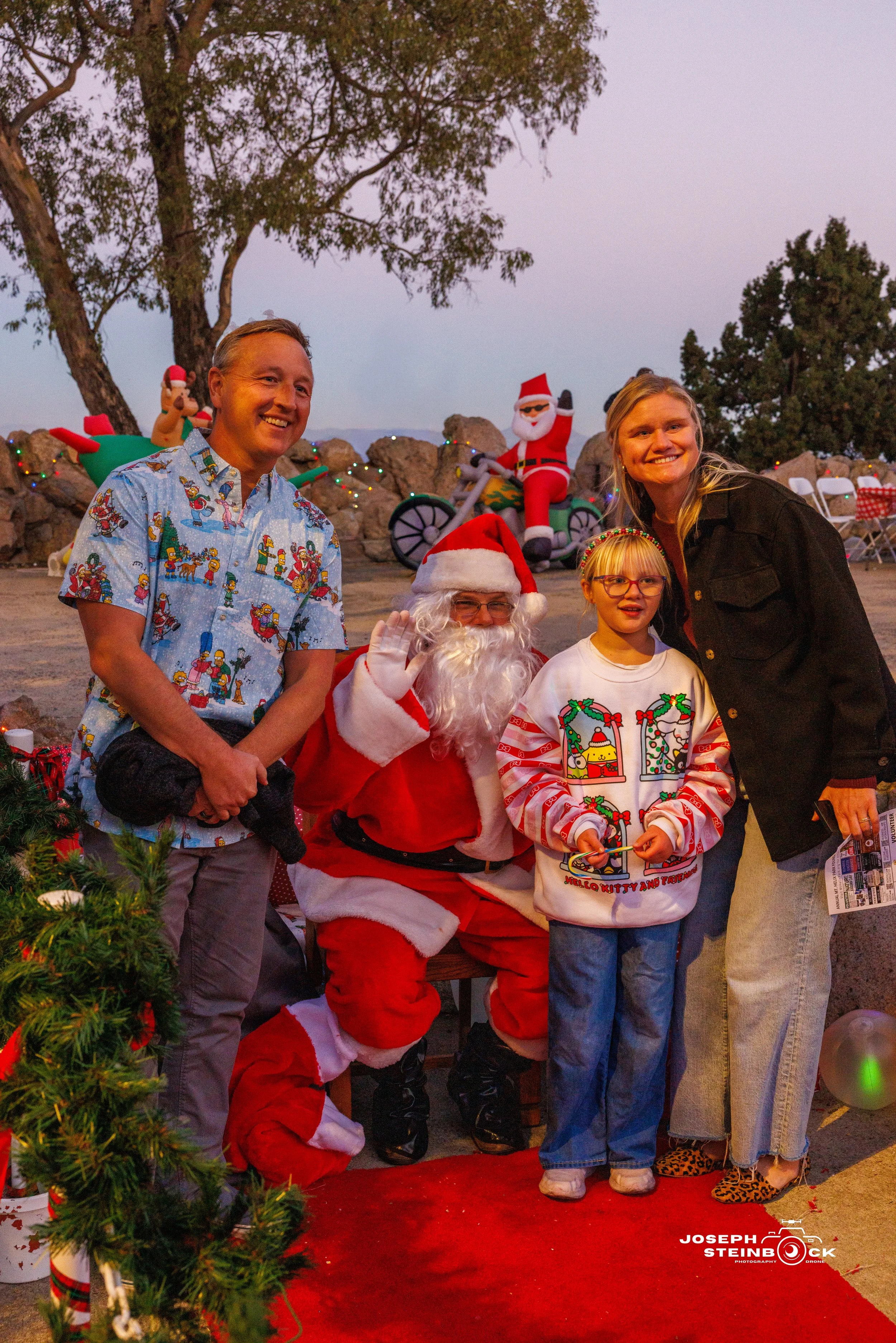 People taking a photo with a person dressed as Santa Claus during a Christmas event outside in the evening with Christmas decorations and a Santa figure on a bike in the background.