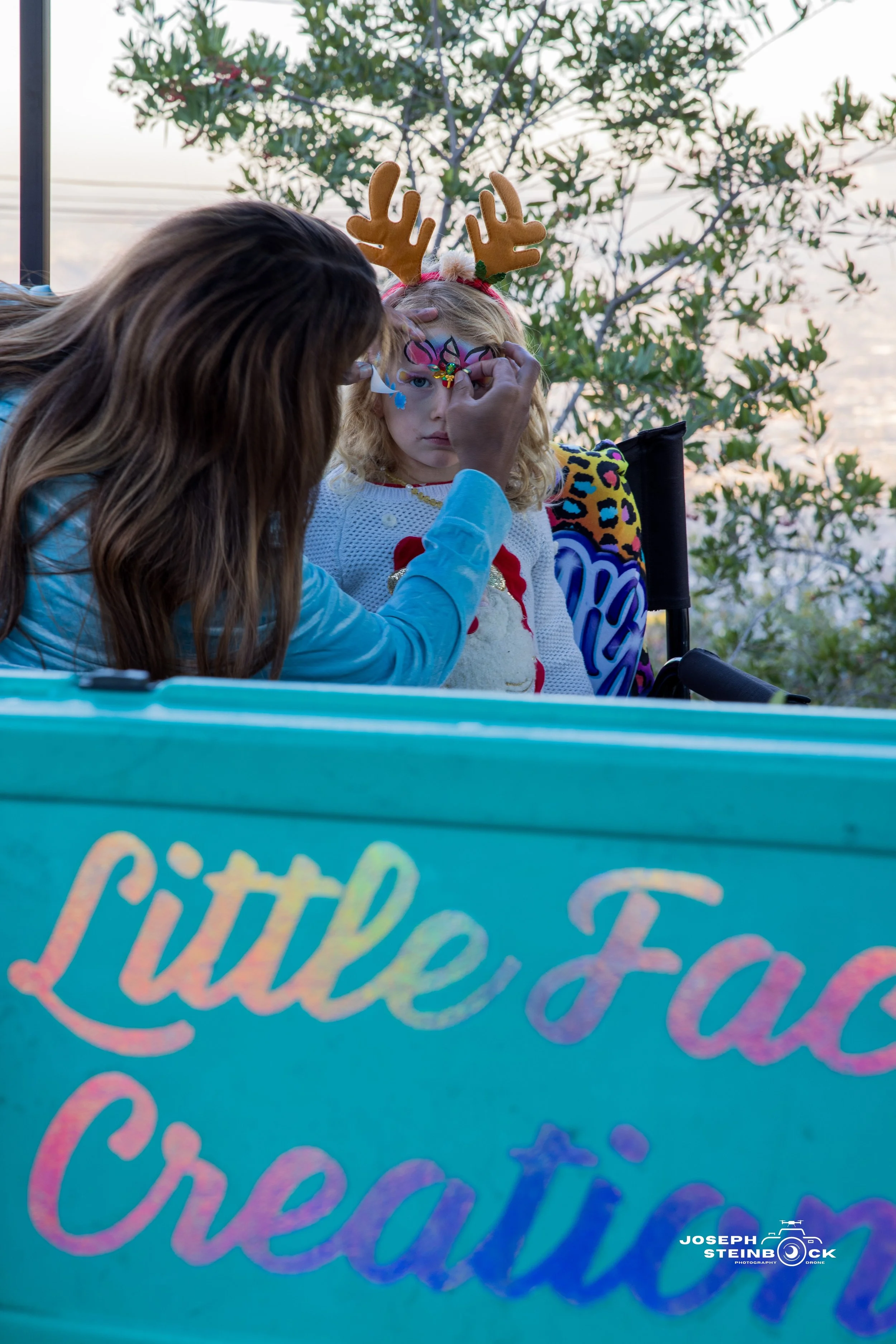 A woman paints a festive face design on a young girl with blonde curly hair, who is wearing reindeer antlers and a colorful sweater. The girl is sitting on a decorated float or booth with the words 'Little Fat Creati' visible on the side, outdoors ne