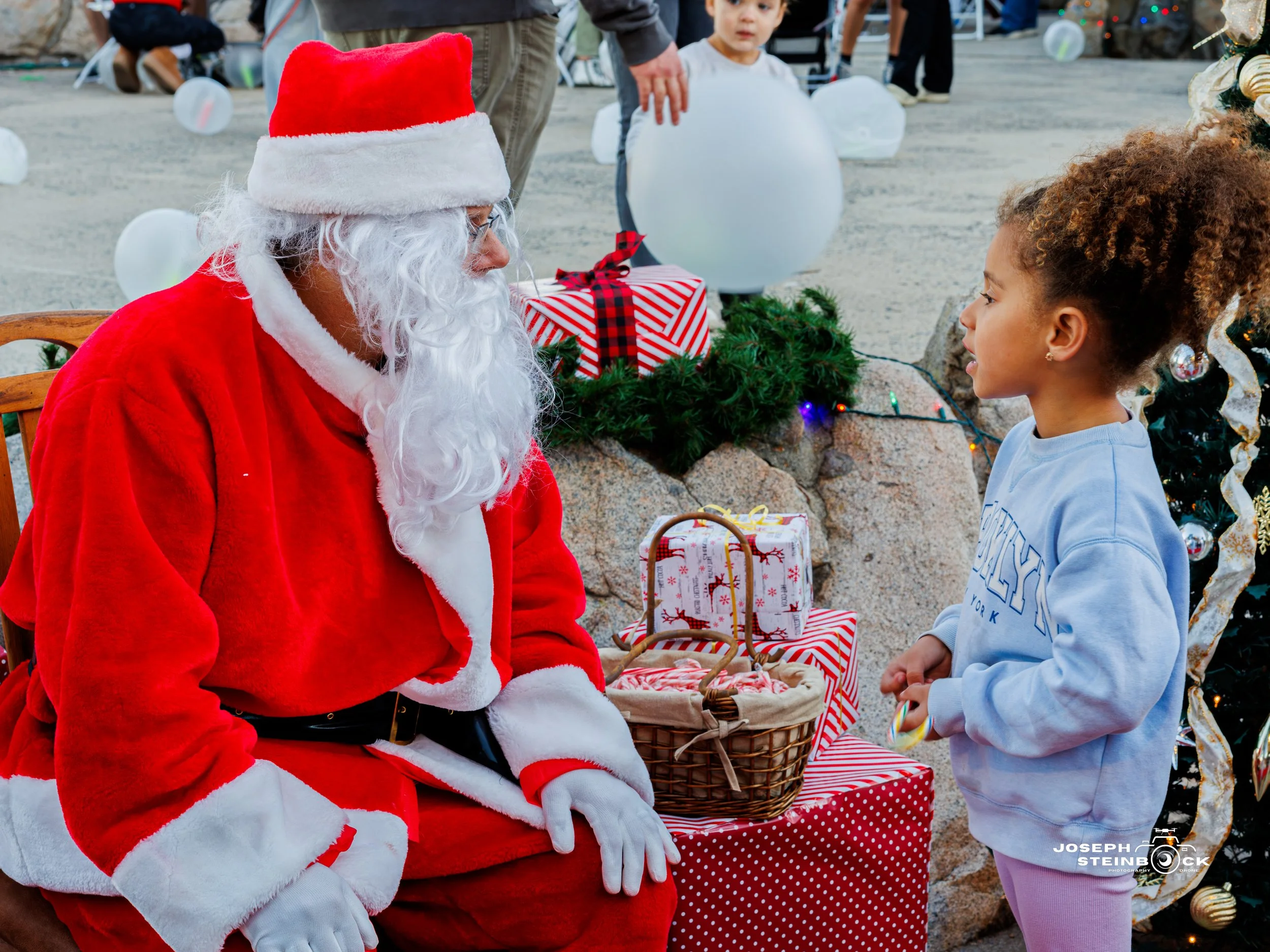 A man dressed as Santa Claus sitting on a wooden chair, talking to a young girl during a Christmas event. The girl is holding a candy cane, and there are wrapped presents, a Christmas tree, and holiday decorations around them.