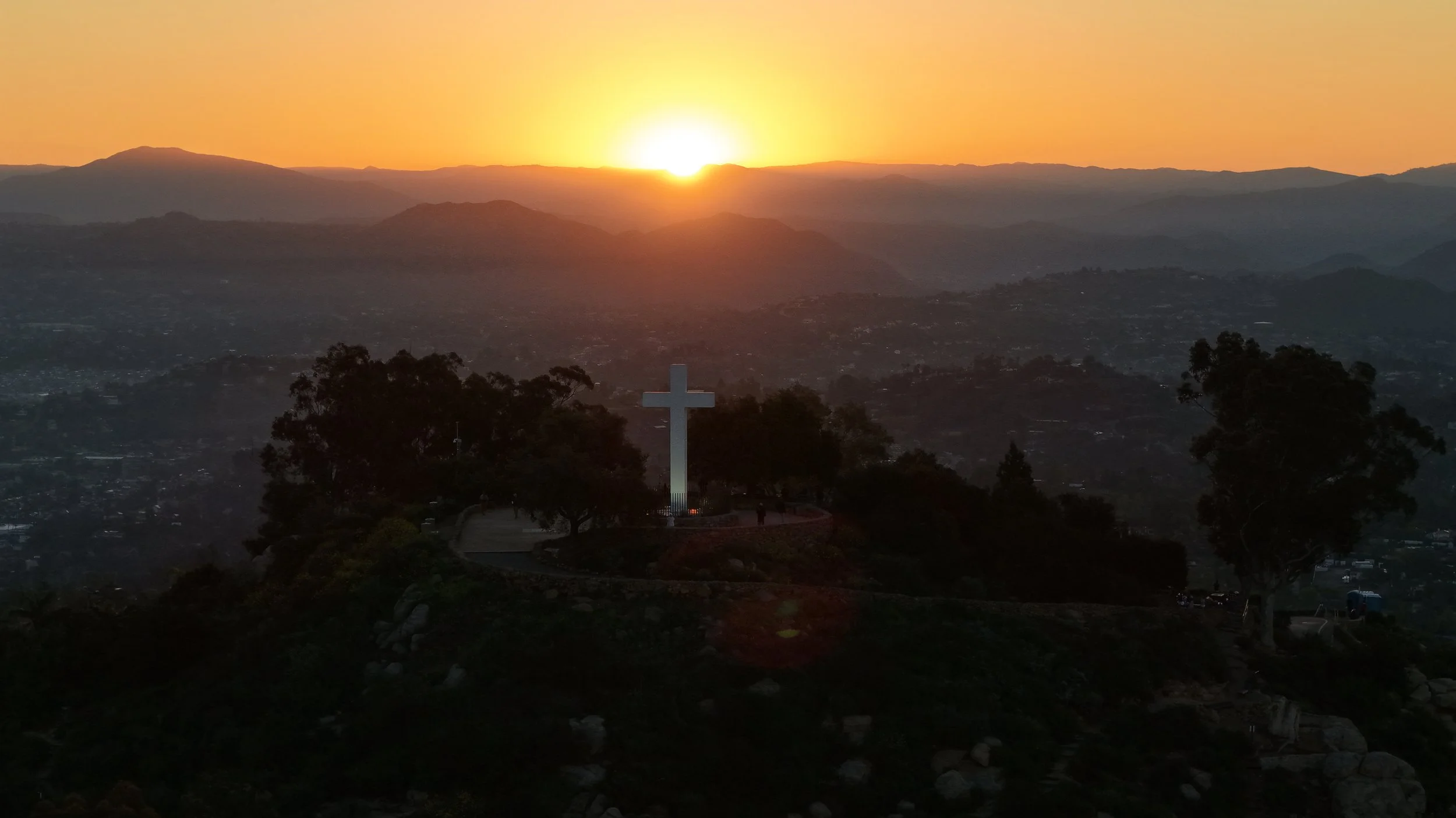 Sunset behind mountains with a large white cross on a hill in the foreground, trees around it, and a city below.