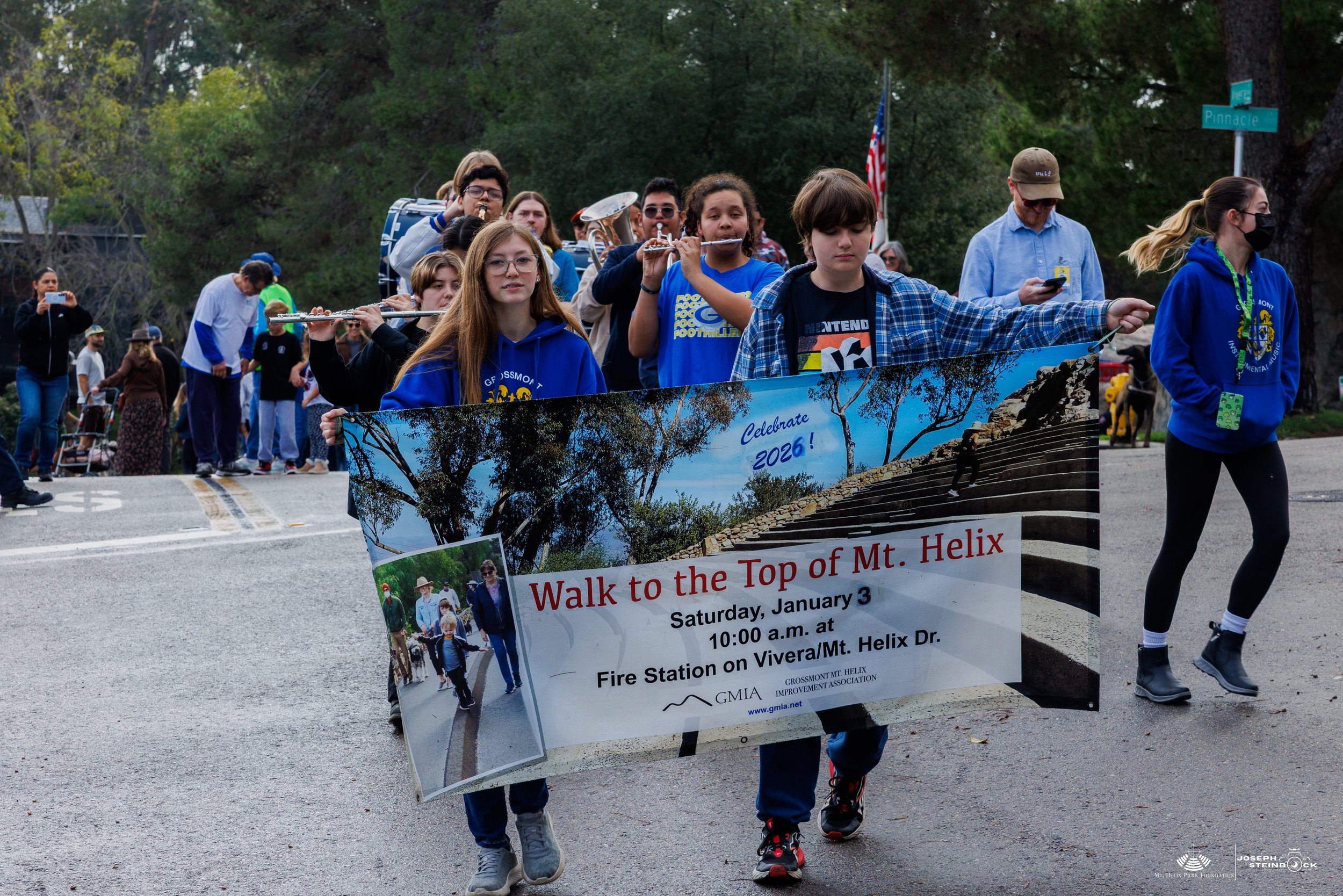 Group of people participating in a walk to the top of Mt. Helix, carrying a banner that reads 'Walk to the Top of Mt. Helix' with details of the event on January 3rd at 10 a.m., near a fire station, with trees and street signs in the background.
