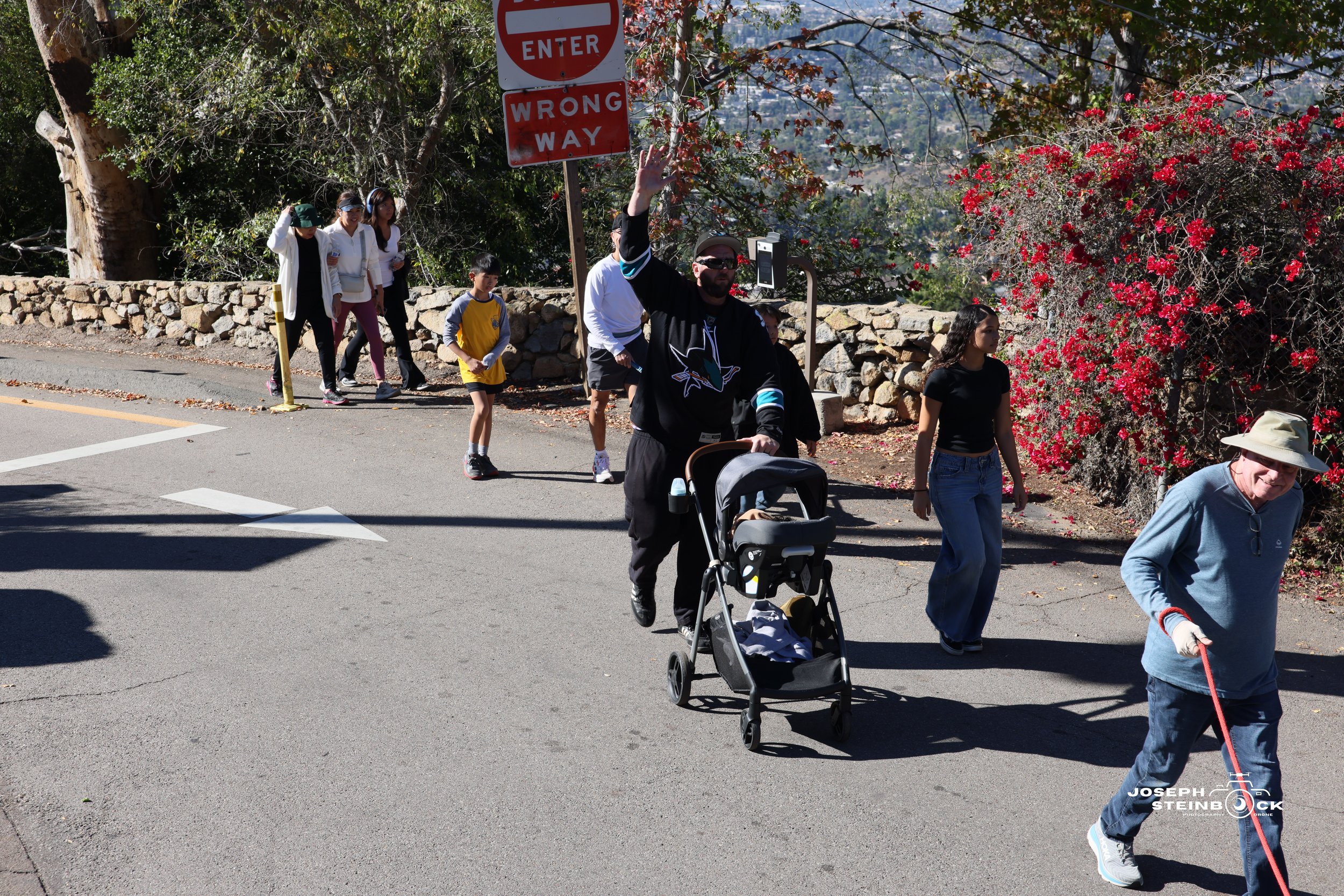 A group of people, including children and adults, are walking across a street. One man is pushing a stroller and waving. There are trees, bushes with red flowers, and a stone wall in the background. Some signs indicating 'Enter' and 'Wrong Way' are v
