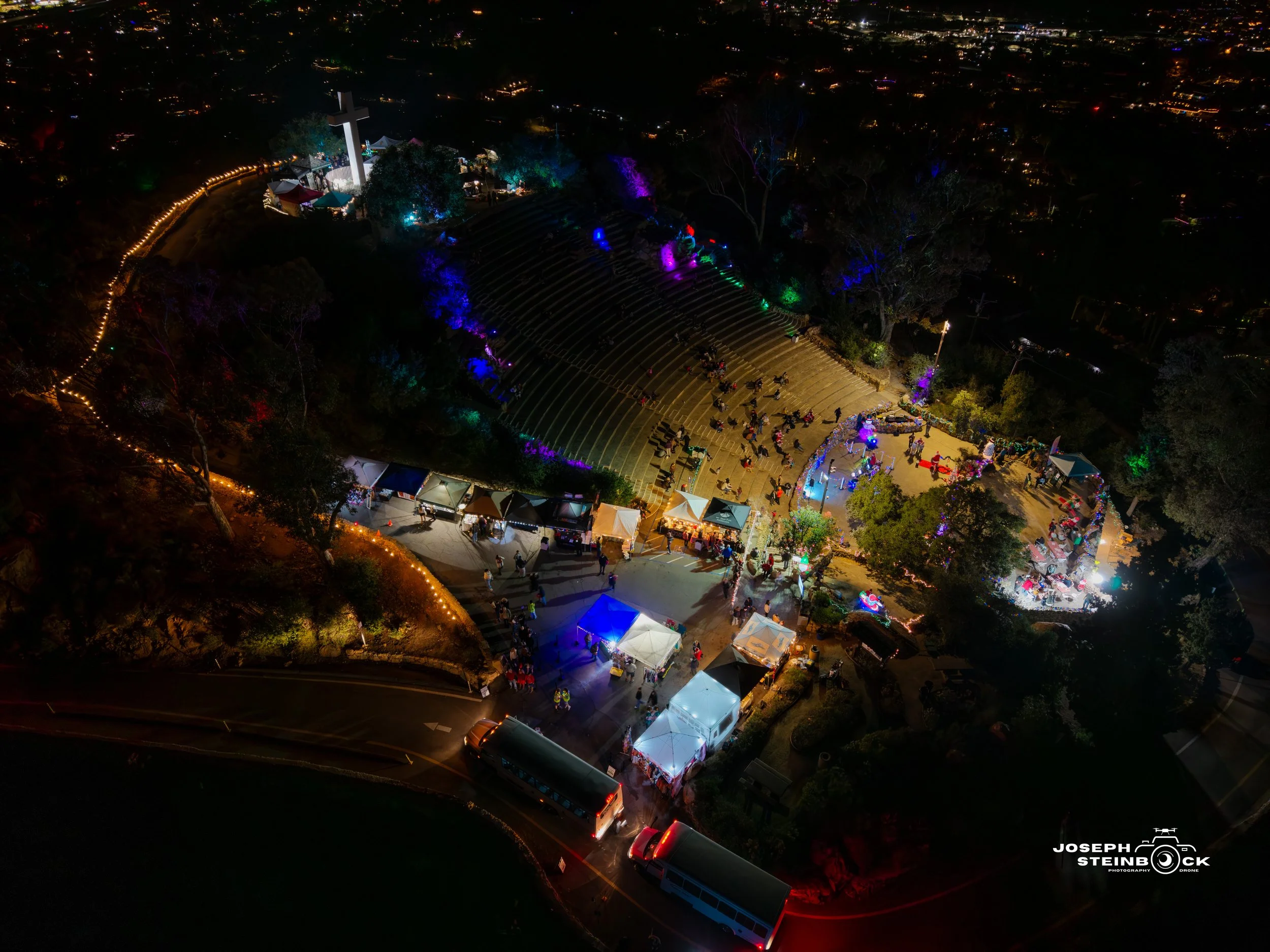An aerial night view of a hilltop outdoor event with a large cross illuminated in white at the top. The area is decorated with colorful lights, tents, and groups of people gathered around, with some on an amphitheater-style seating area.