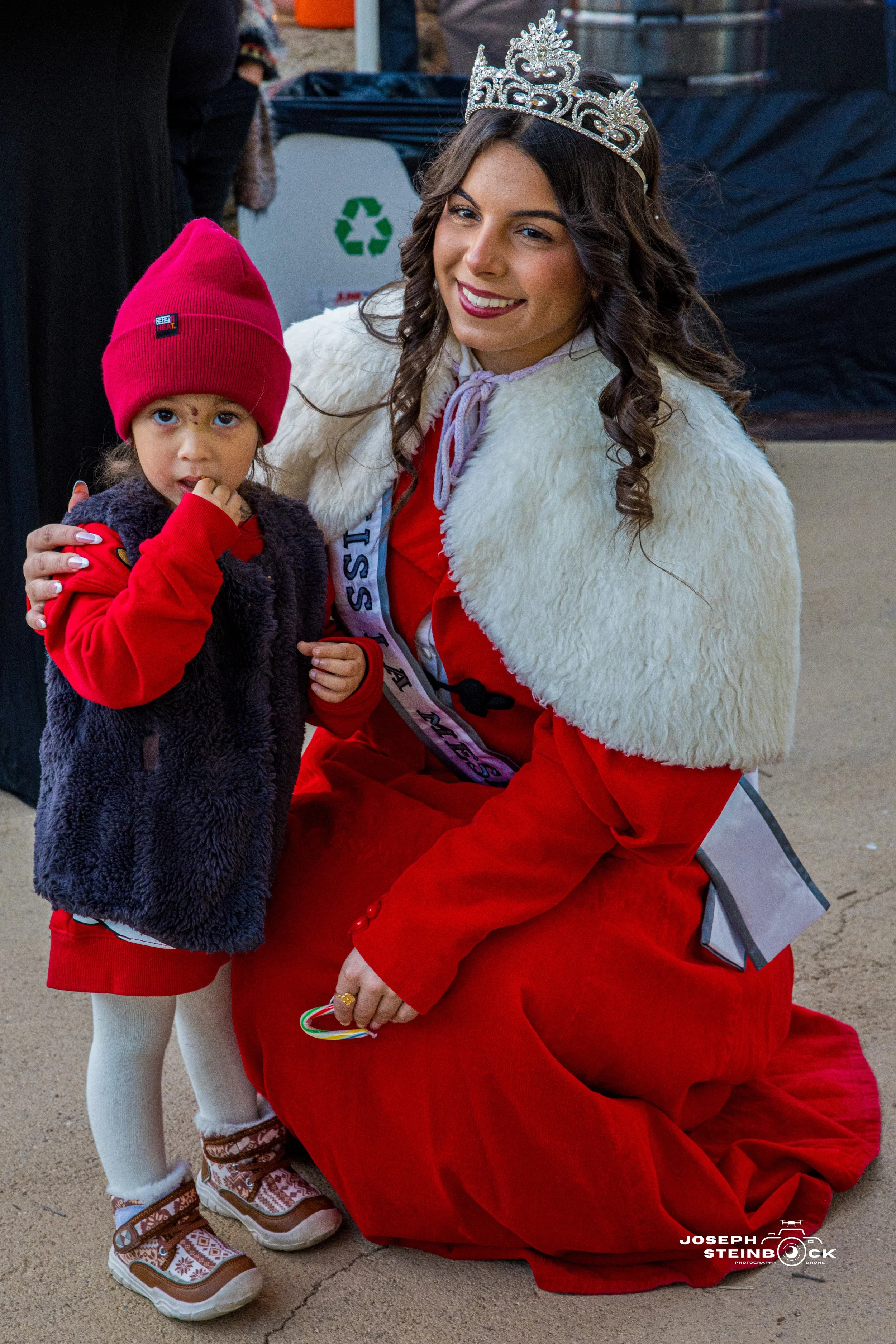 A woman dressed as Miss Santa with a sash and tiara kneeling next to a girl in a red hat and festive winter attire at a holiday event.