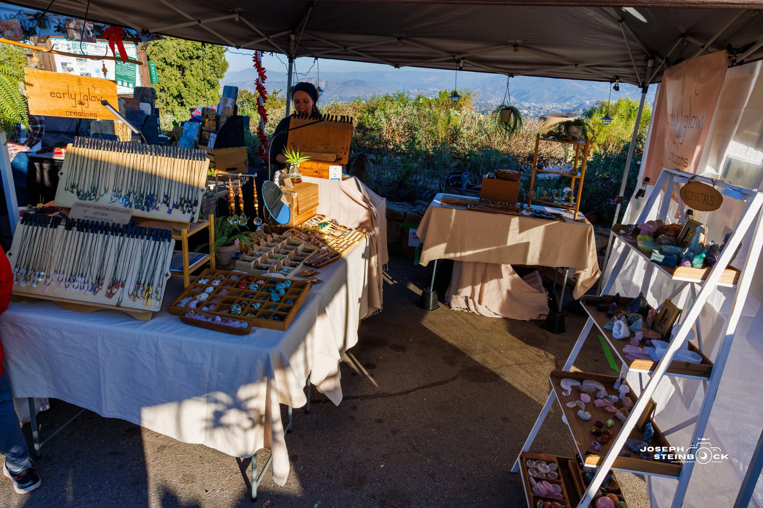 Outdoor craft market stand with jewelry and gemstone displays under a canopy, with a woman arranging items, in a scenic setting with mountains and trees in the background.