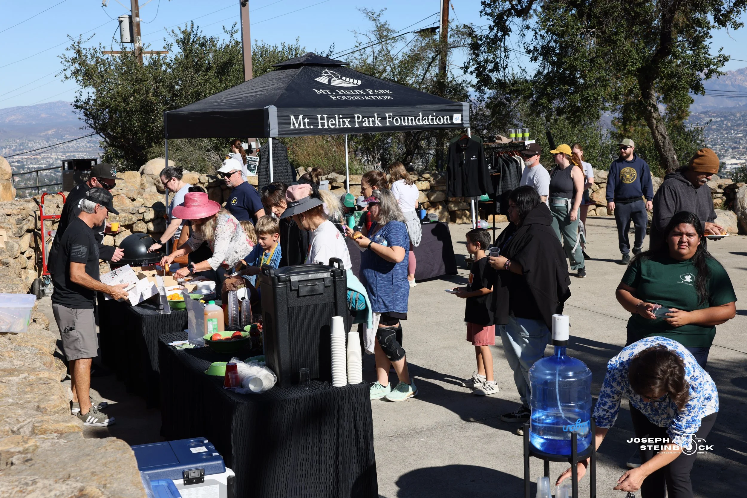 People gather around a catering table with a black tablecloth at an outdoor event under a canopy that reads "Mt. Helix Park Foundation". Some are drinking, walking, or serving food, with tents and trees in the background.