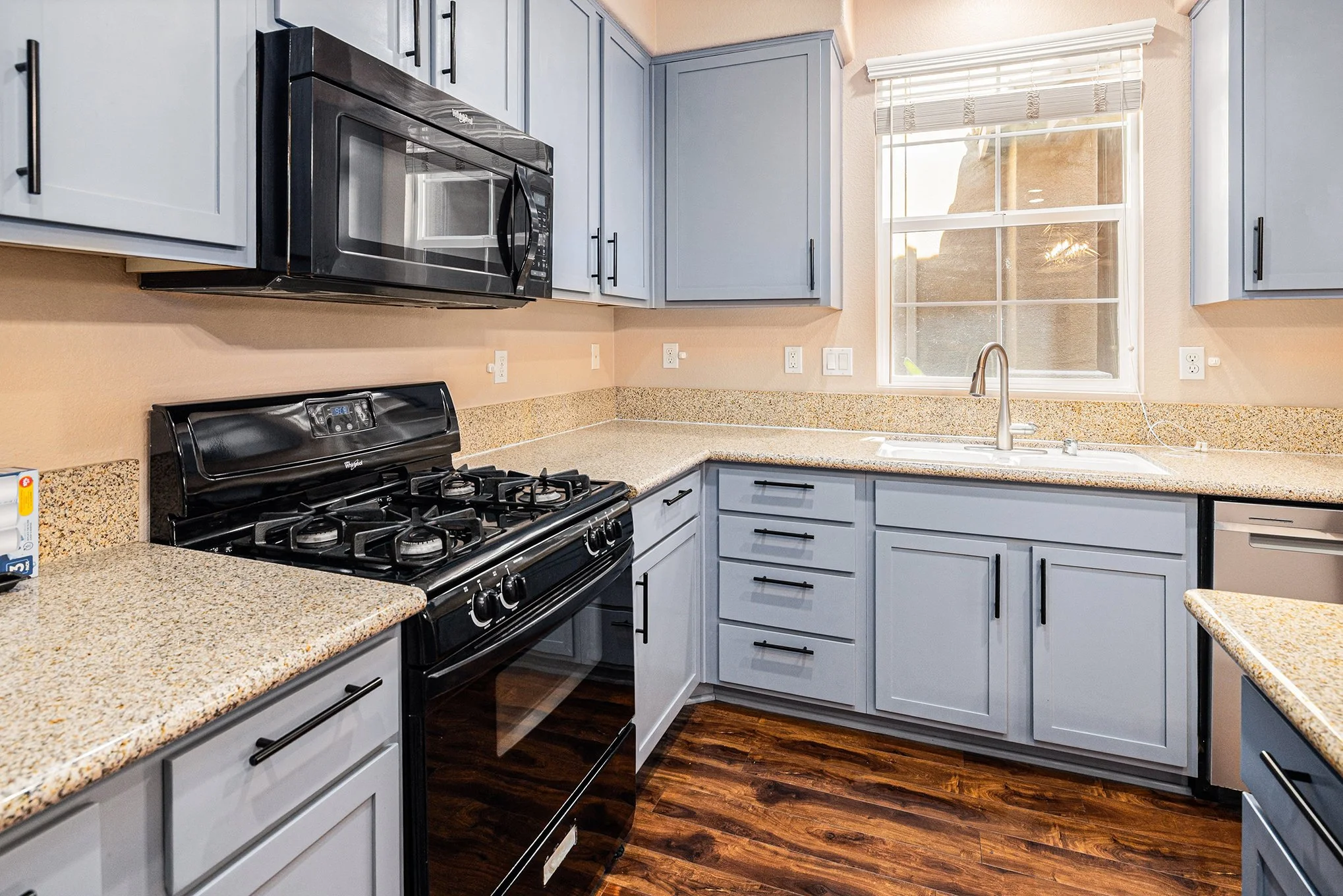 Kitchen with light blue cabinets, black stove and microwave, beige countertops, a window above the sink, and dark wood flooring.