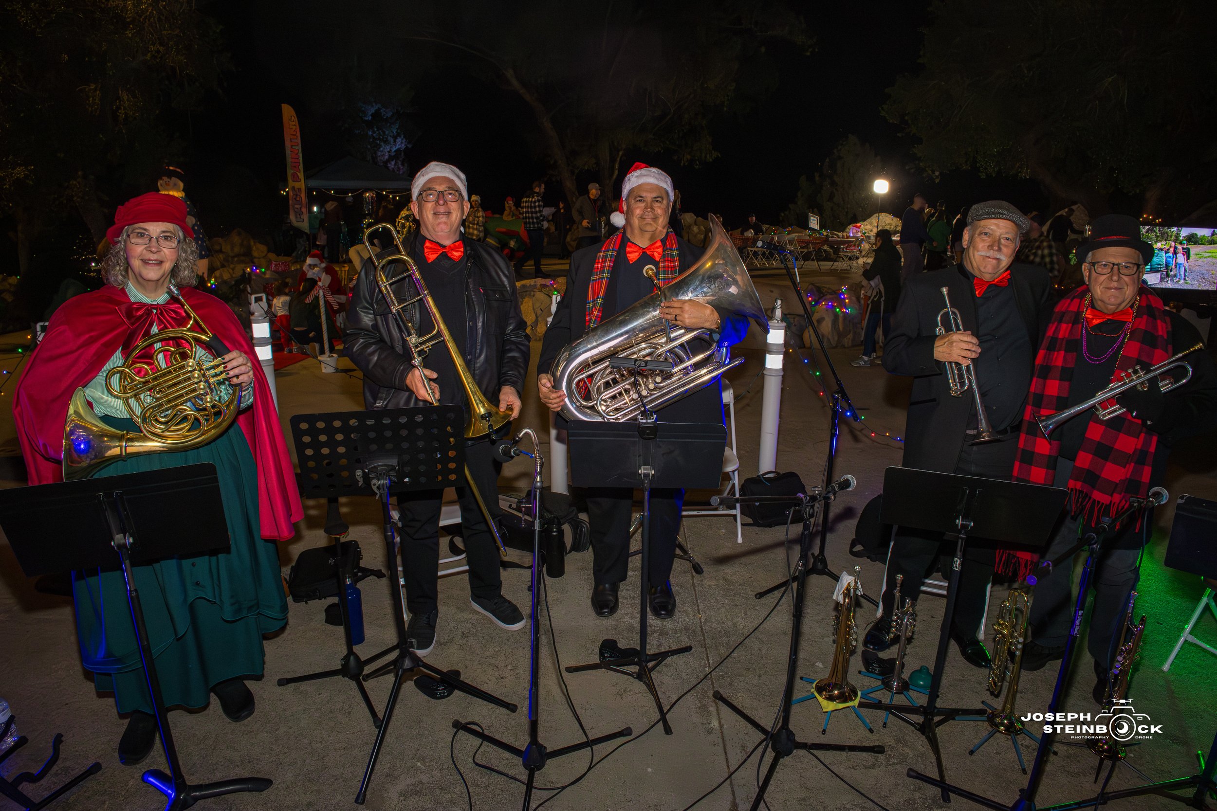 Band of six musicians wearing holiday costumes, playing brass instruments, standing outdoors at night with Christmas lights and decorations in the background.