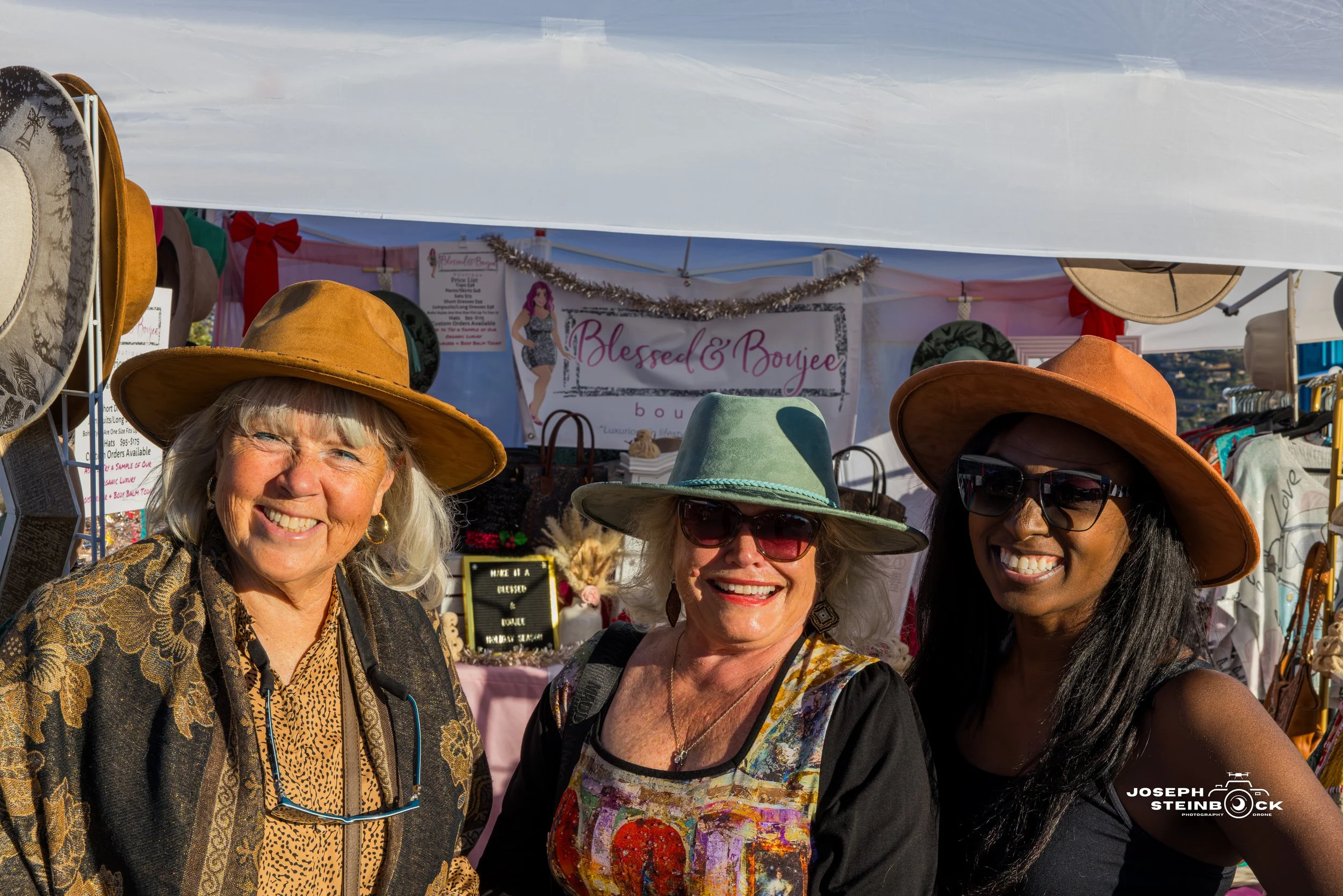 Three women smiling at a market booth, wearing wide-brimmed hats and sunglasses, with jewelry and accessories displayed in the background at the 'Blessed & Boujee' booth.