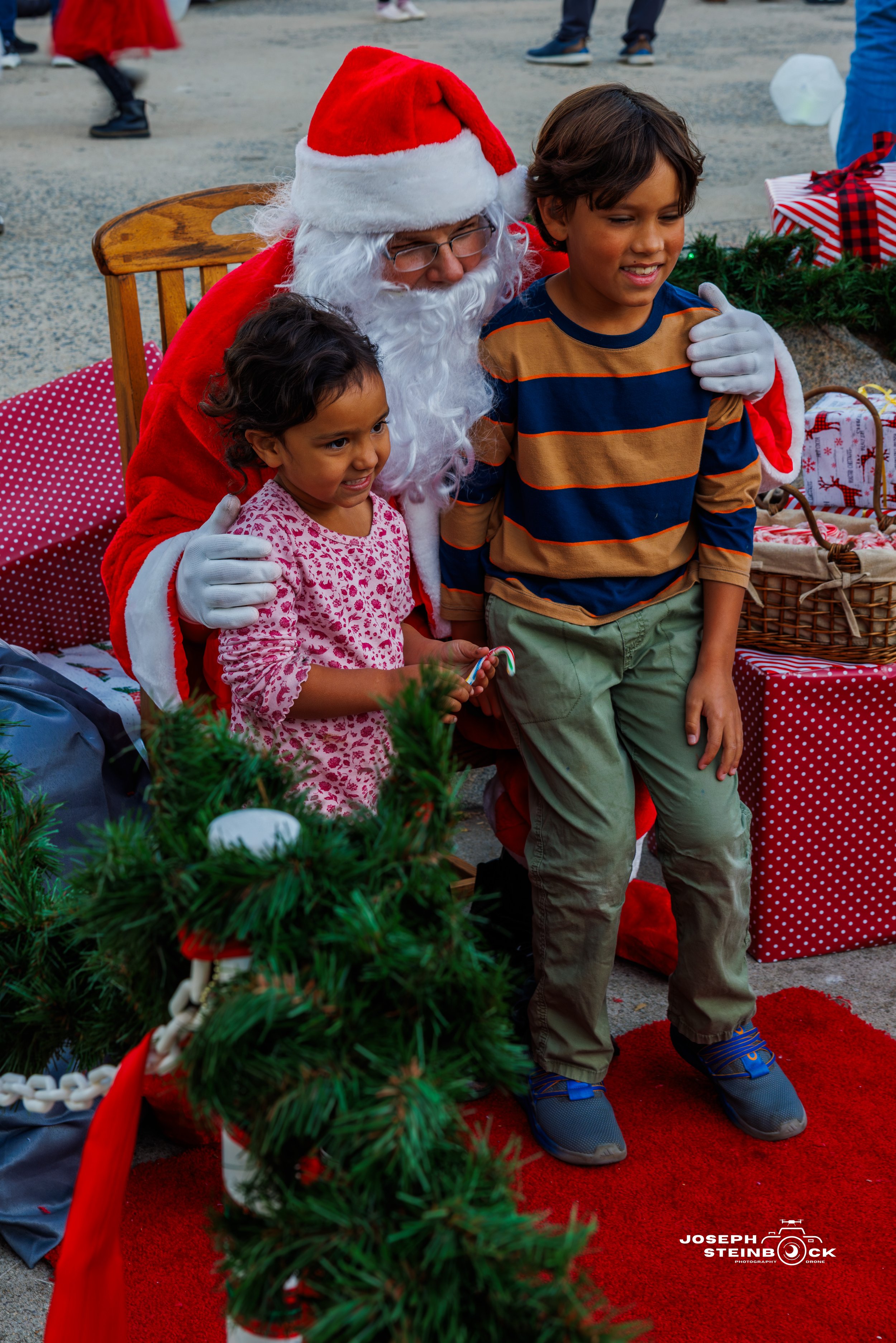 Santa Claus sitting on a wooden chair with a little girl and a boy, hugging each other during a Christmas celebration outdoors, with wrapped presents and greenery around them.